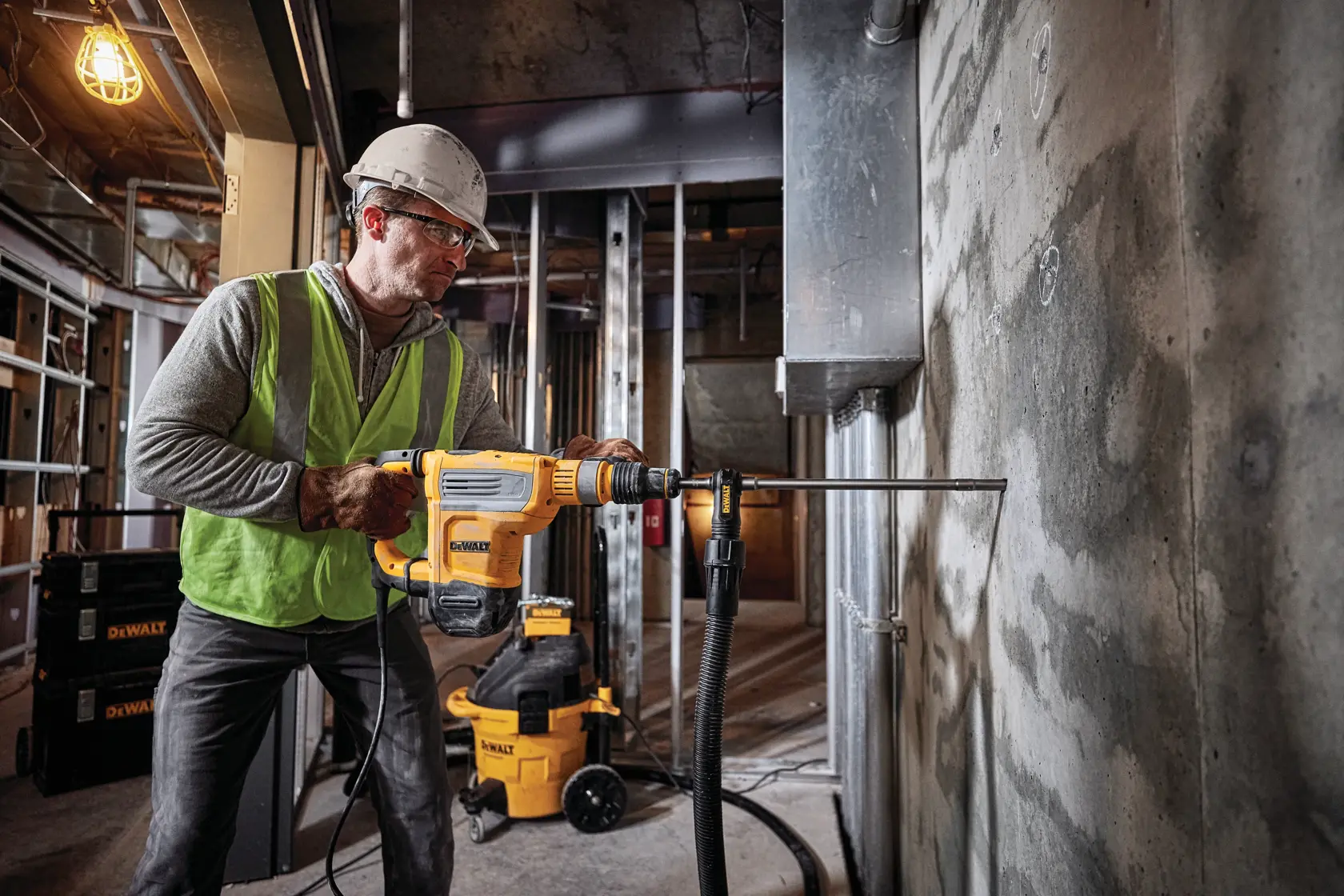 A worker wearing a safety helmet and vest operates a DEWALT rotary hammer drill (SKU: D25614K) to drill into a concrete wall at a construction site. DEWALT equipment and tools are visible in the background.