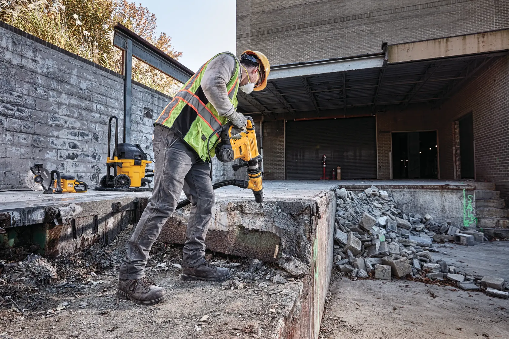 A construction worker wearing safety gear and a reflective vest uses a DEWALT demolition hammer to break up concrete at an outdoor job site. Additional DEWALT tools and equipment are visible in the background.