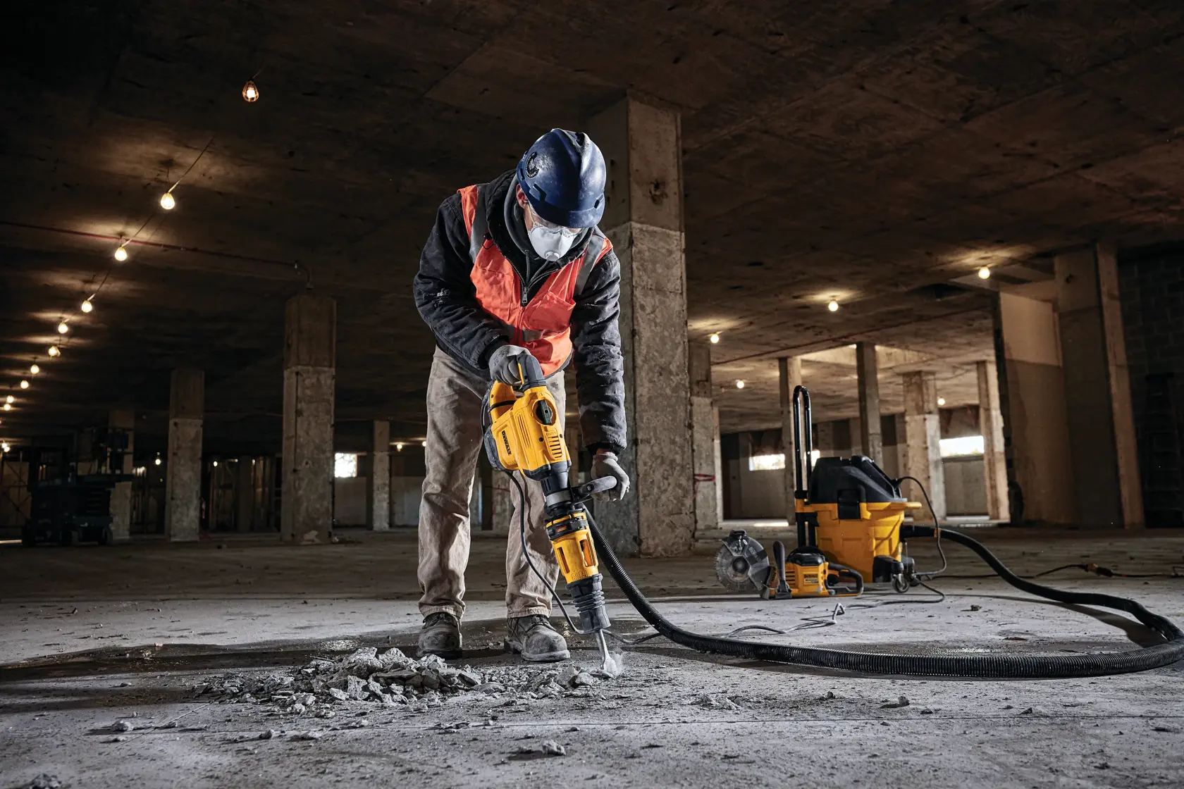 A worker wearing safety gear operates a DEWALT demolition hammer (SKU: D25832K) to break concrete on the floor of a large, unfinished building. A yellow industrial vacuum cleaner is connected to the tool via a hose, visible in the background.