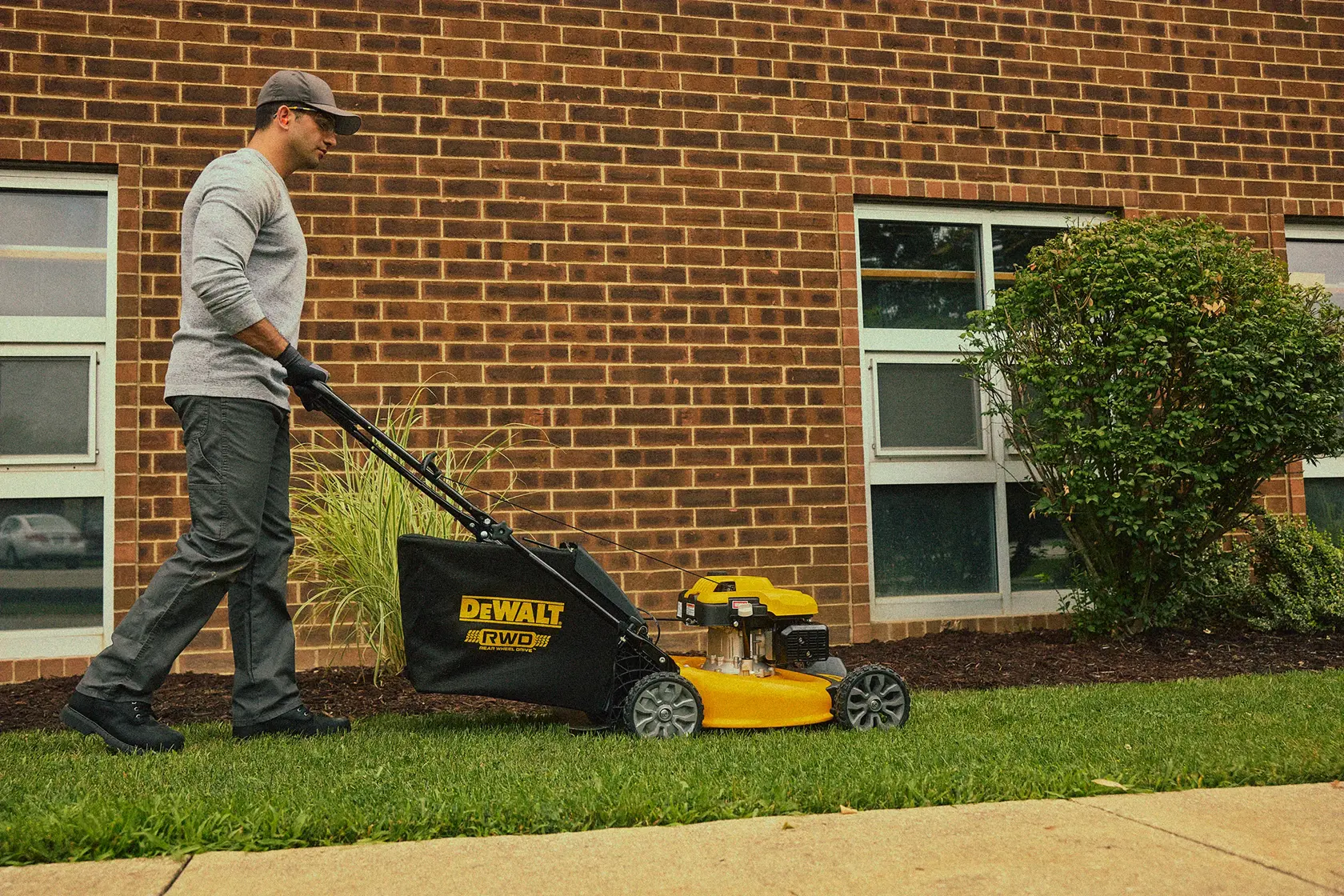 A person pushes a yellow DEWALT lawn mower on a grassy area next to a brick building with windows and bushes.