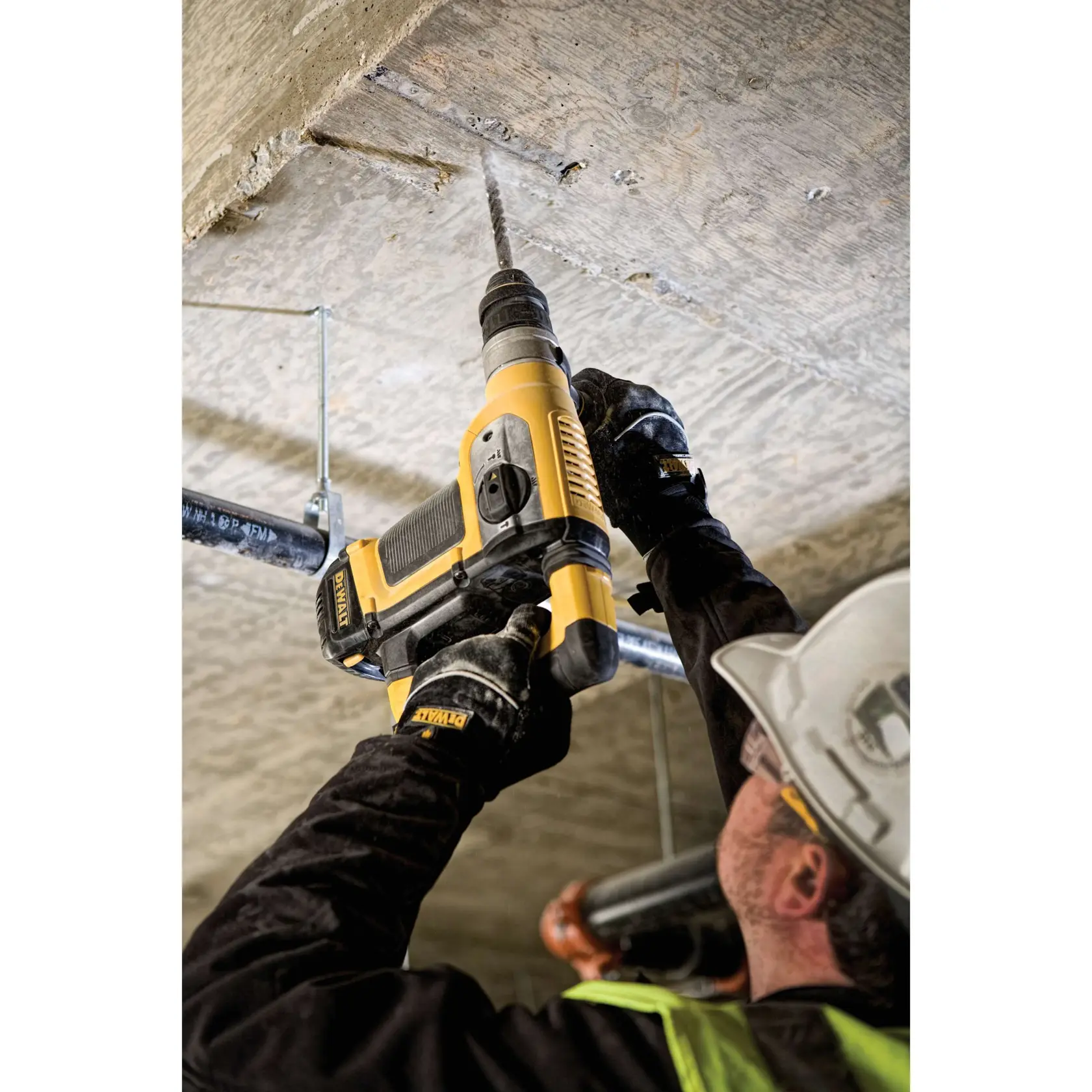 A person wearing protective gear uses a yellow DEWALT rotary hammer drill to drill into a concrete ceiling at a construction site.