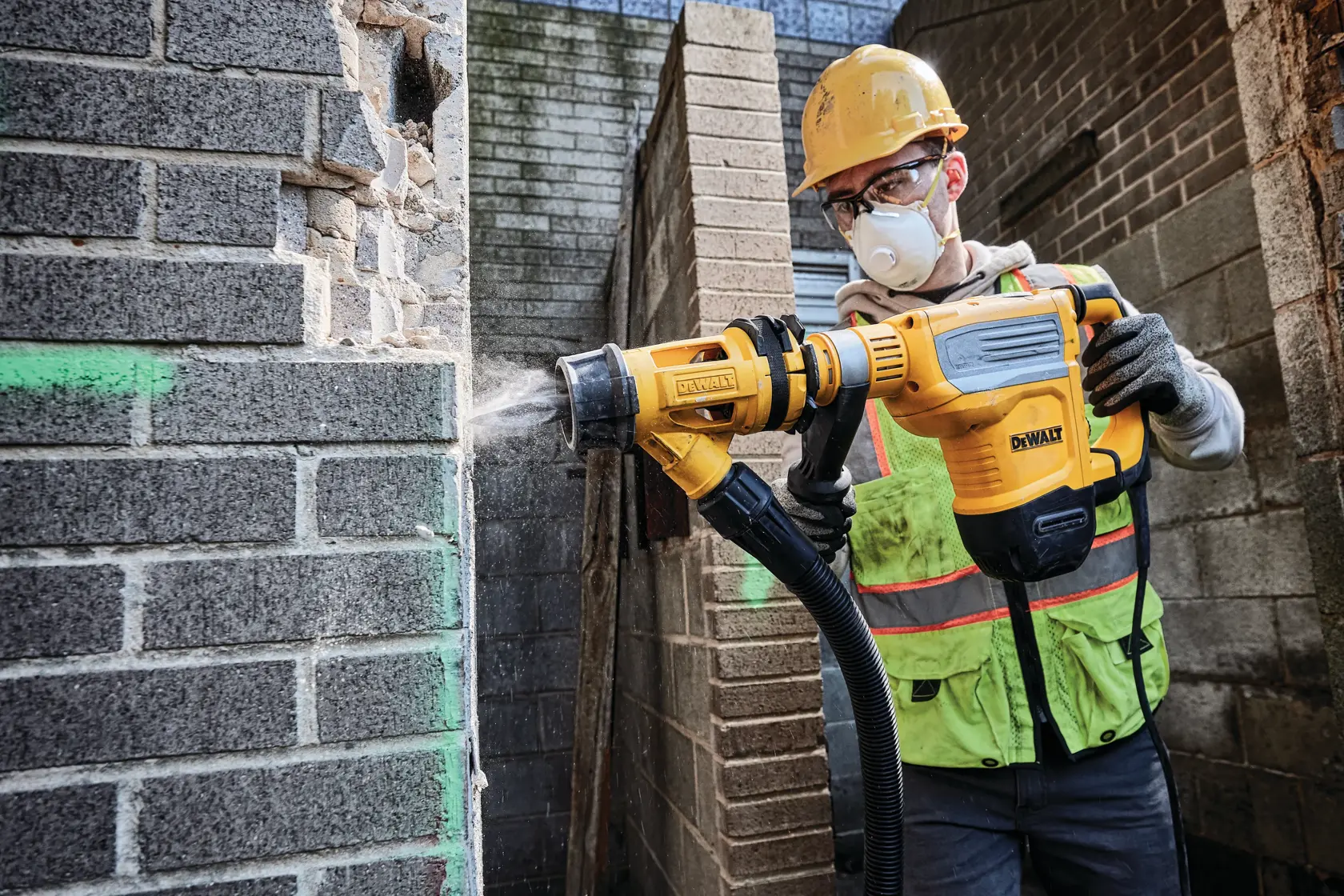 A construction worker wearing a safety helmet and vest operates a yellow DEWALT D25614K demolition hammer drill with dust extraction on a brick wall.