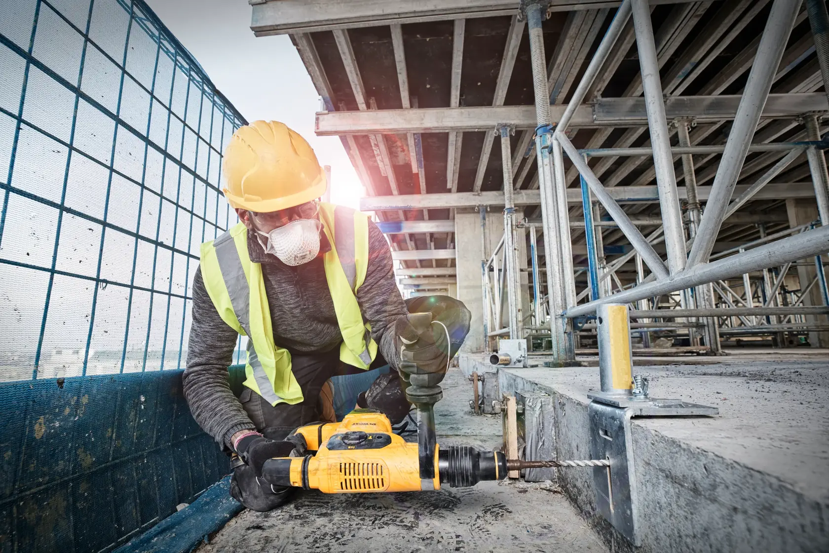 A construction worker wearing a yellow hard hat and reflective vest is using a yellow DEWALT hammer drill to drill into a concrete surface at a building site.