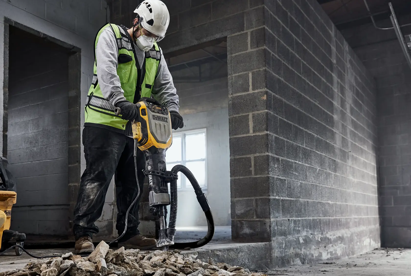 A construction worker wearing a safety helmet and high-visibility vest operates a DEWALT D25966K demolition hammer, breaking concrete on a construction site. The worker stands among rubble with the tool connected to a vacuum hose for dust extraction.