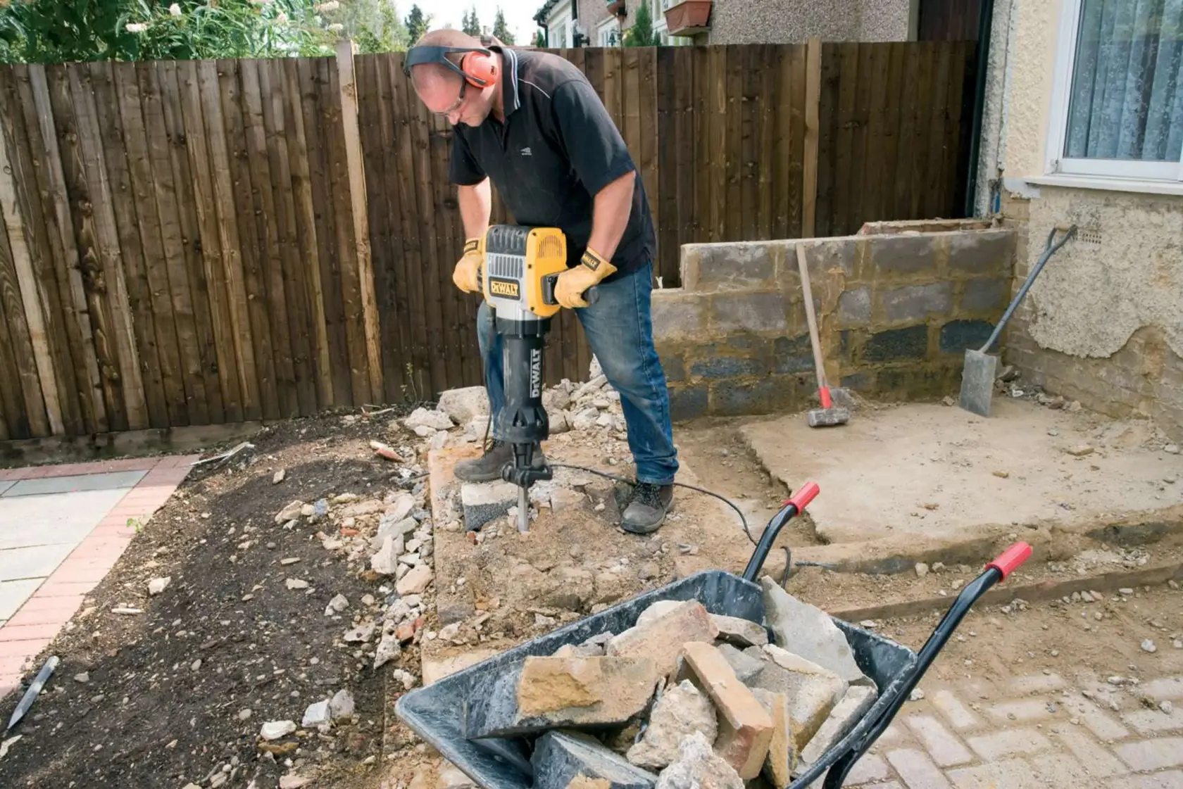 A person using a DEWALT electric demolition hammer (model D25980) to break concrete outdoors, with a wheelbarrow full of rubble and stones in the foreground.