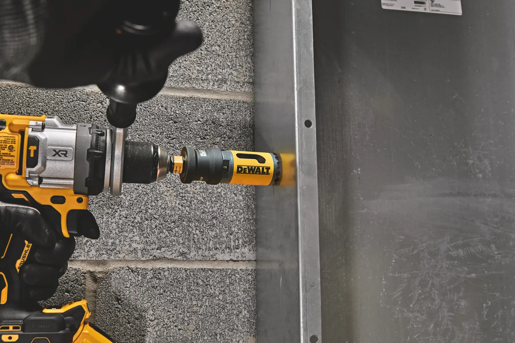 DEWALT drill with hole saw attachment drilling into a metal surface beside a cinder block wall.