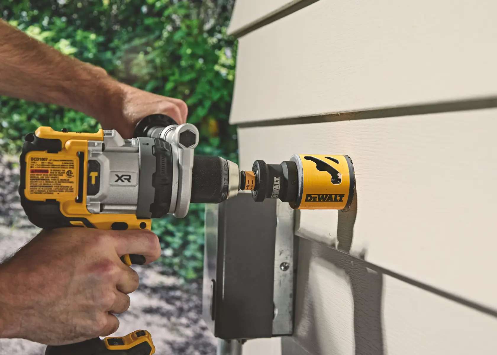 Hand using DEWALT drill with hole saw attachment on exterior wall siding.