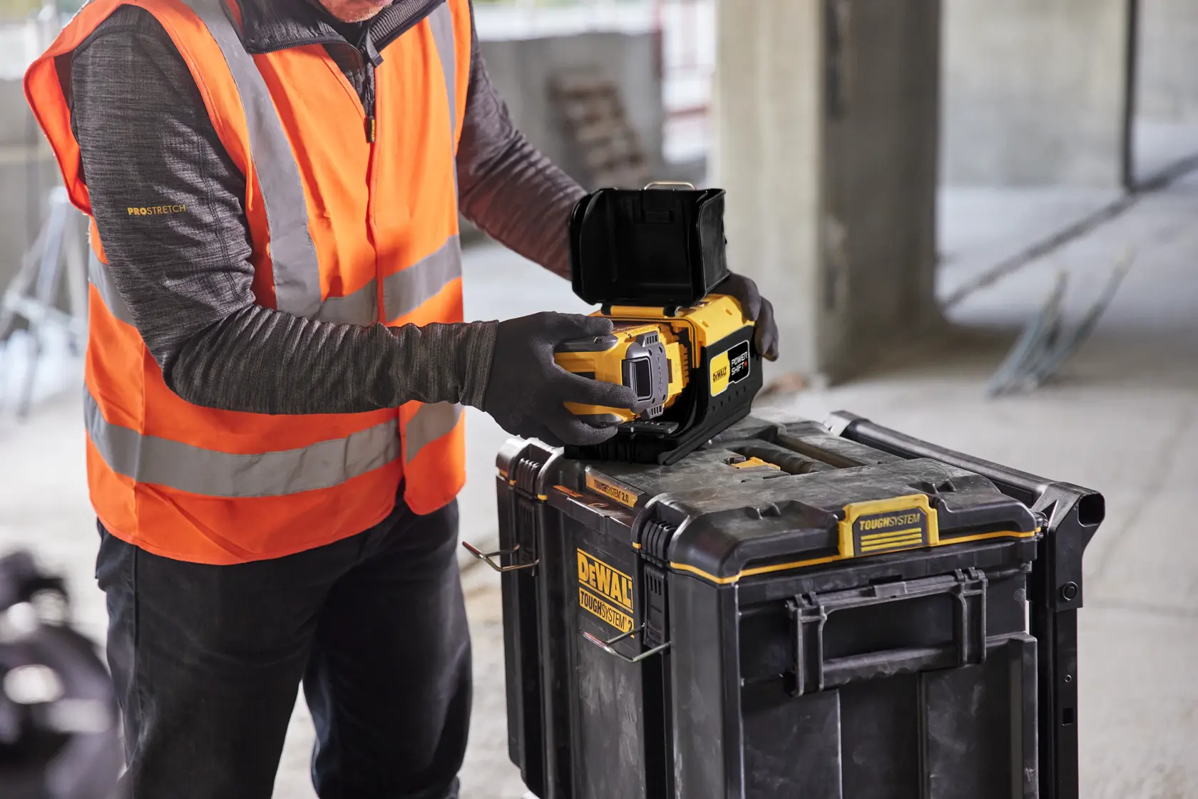 A person wearing an orange safety vest and gloves is placing a DEWALT camera or photography device on top of a DEWALT storage box at a construction site.