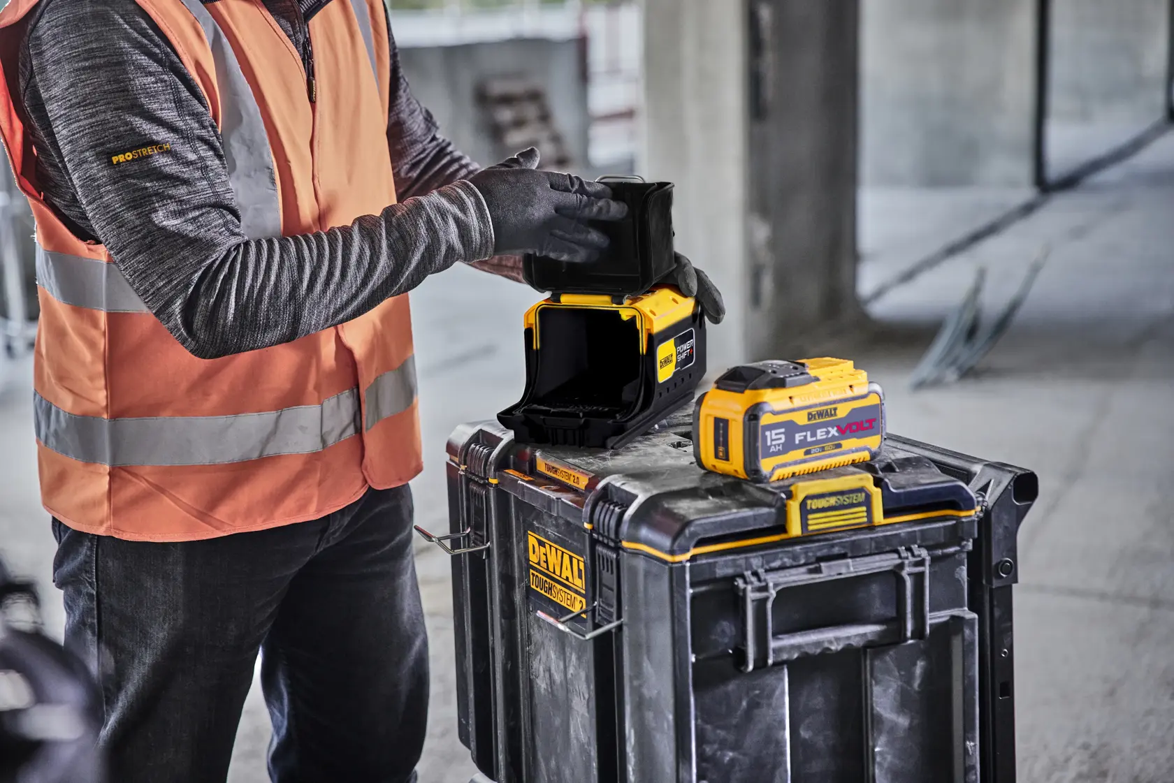 A person wearing an orange safety vest and gloves is handling a DEWALT camera device placed on top of a black DEWALT ToughSystem toolbox. Another yellow DEWALT FLEXVOLT battery is also visible on the toolbox, and the scene appears to be set at a construction site.