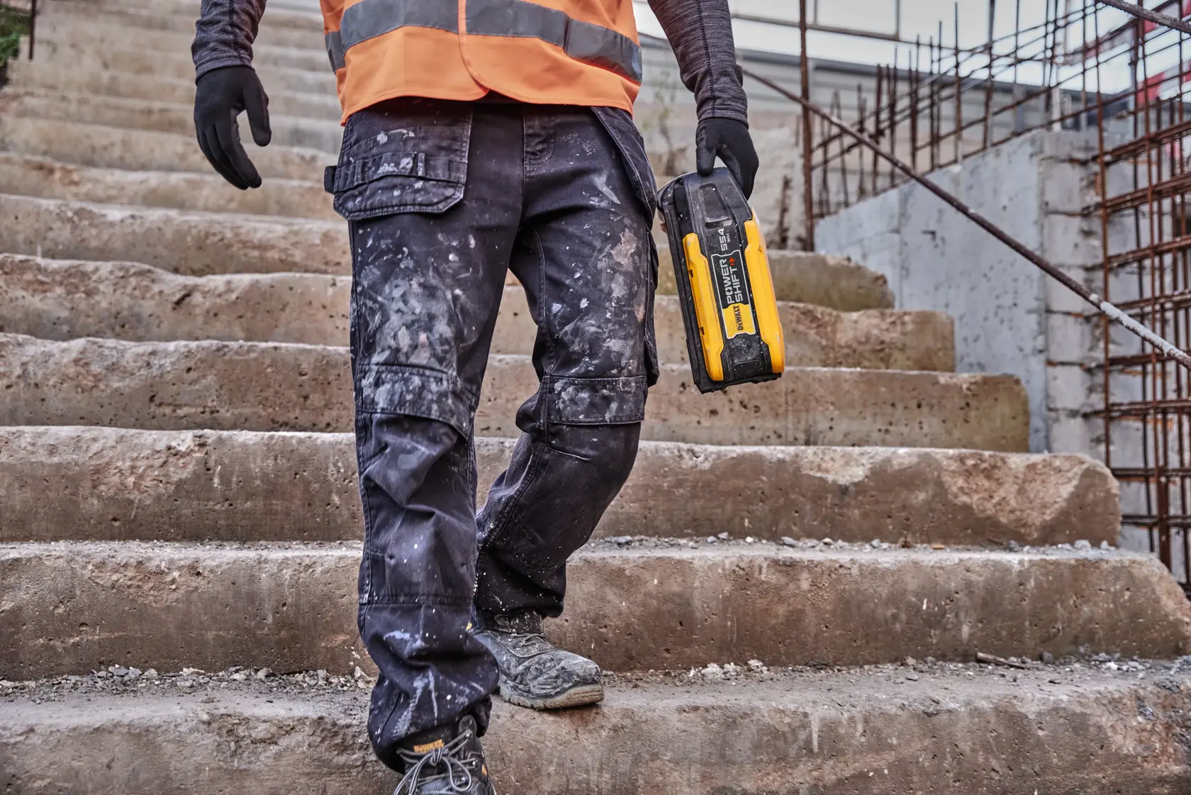 A person wearing work gloves, a high-visibility orange vest, and paint-splattered cargo pants walks down concrete steps at a construction site, holding a DEWALT portable power tool battery.