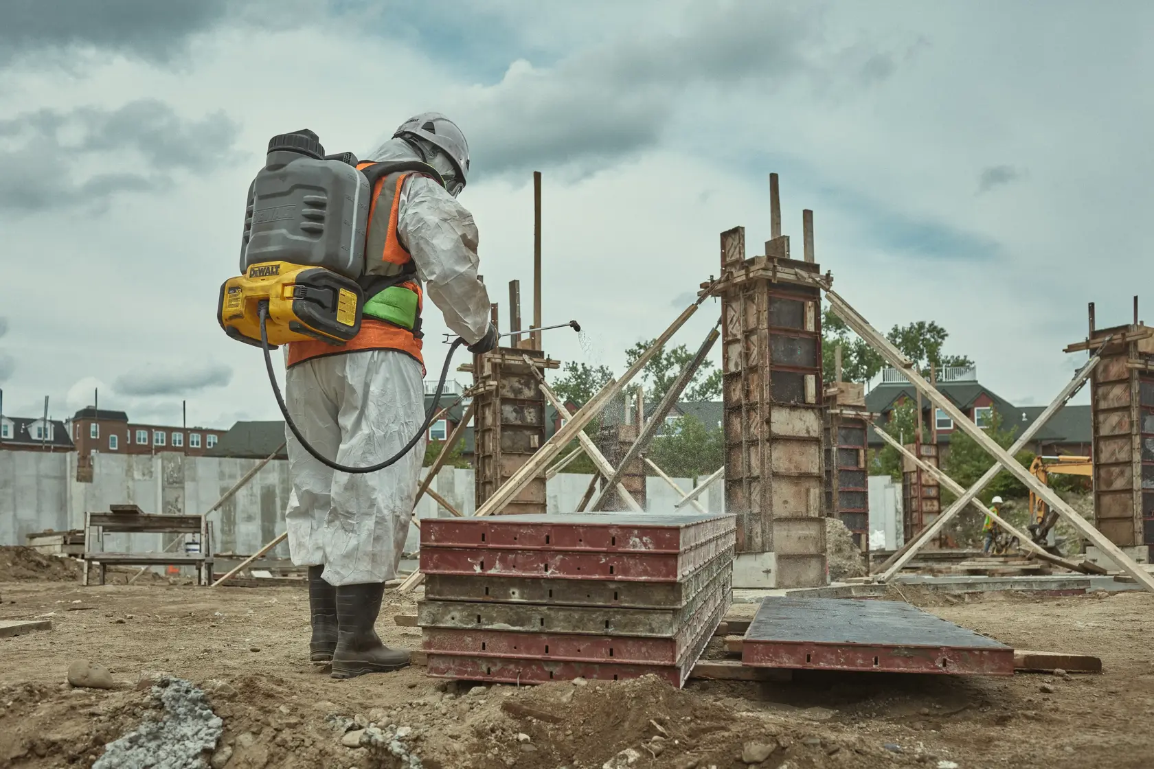 A person wearing protective gear and a DEWALT backpack sprayer is working at a construction site with concrete pillars and wooden frameworks.