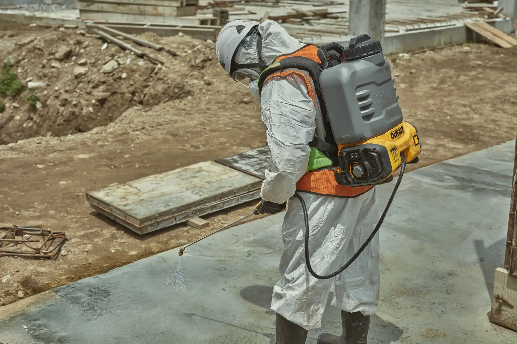 A person wearing protective clothing and a helmet uses a DEWALT backpack sprayer to spray liquid onto a concrete surface at a construction site.