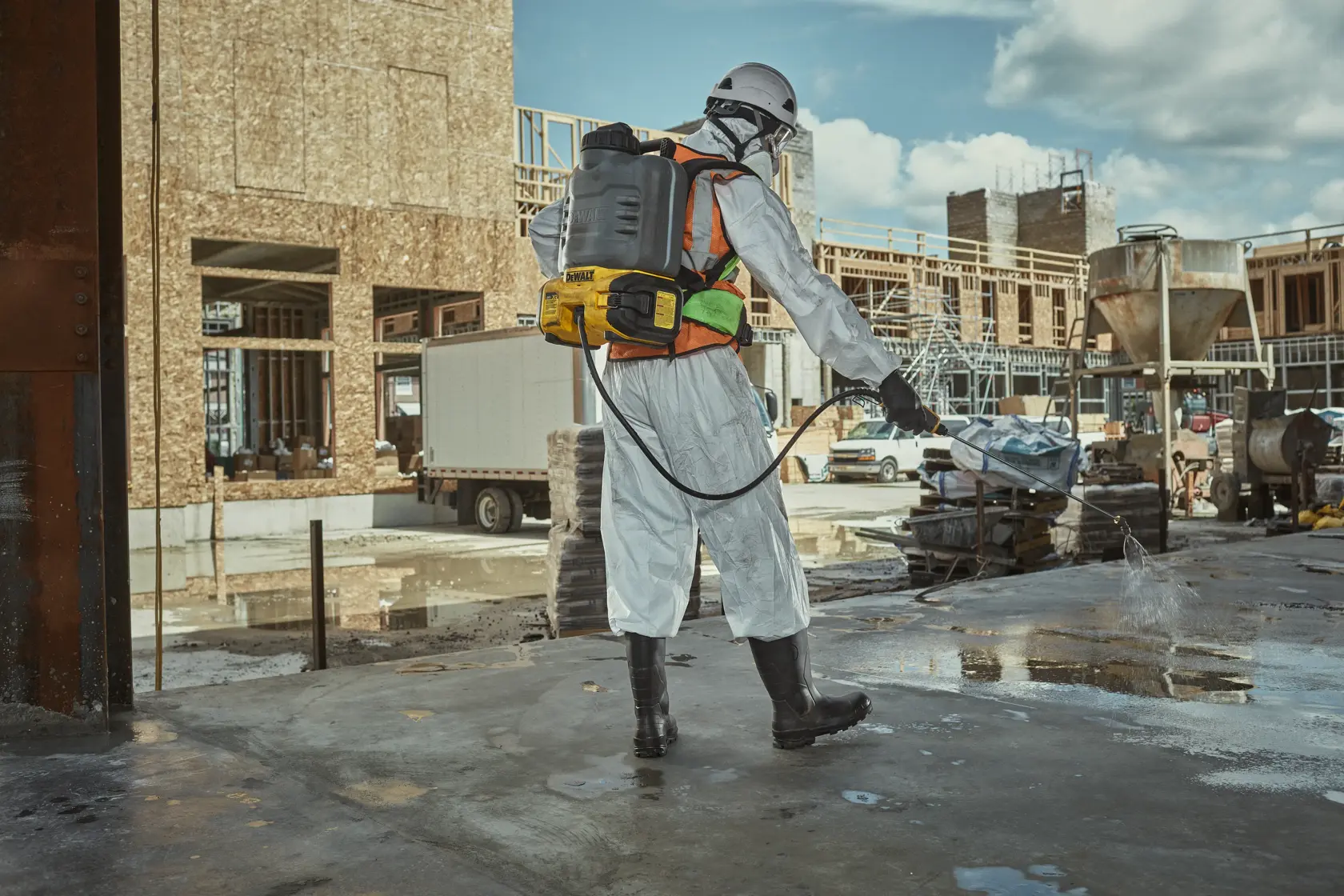 Person wearing safety gear and a DEWALT backpack sprayer is spraying liquid onto the ground at a construction site.
