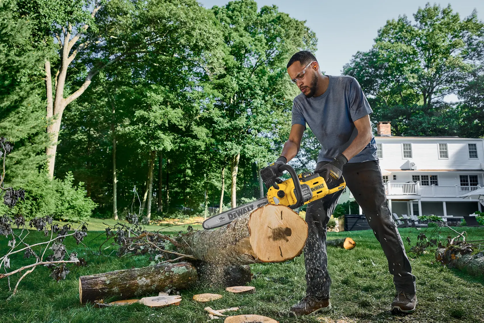 Person uses a DEWALT chainsaw to cut a log outdoors with trees and a house in the background.