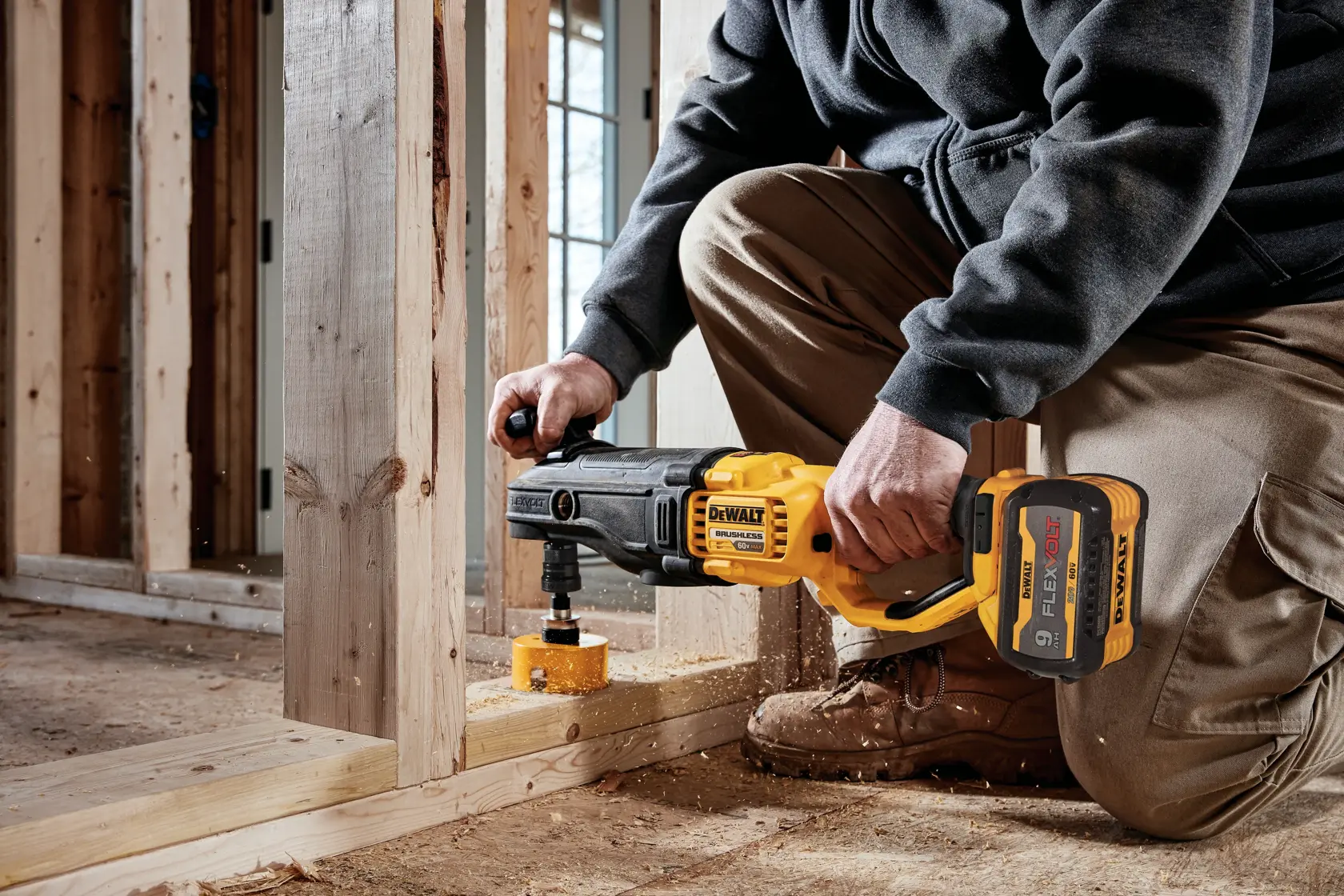 A person using a DEWALT cordless rotary drill with a hole saw attachment to drill through a wooden beam at a construction site.