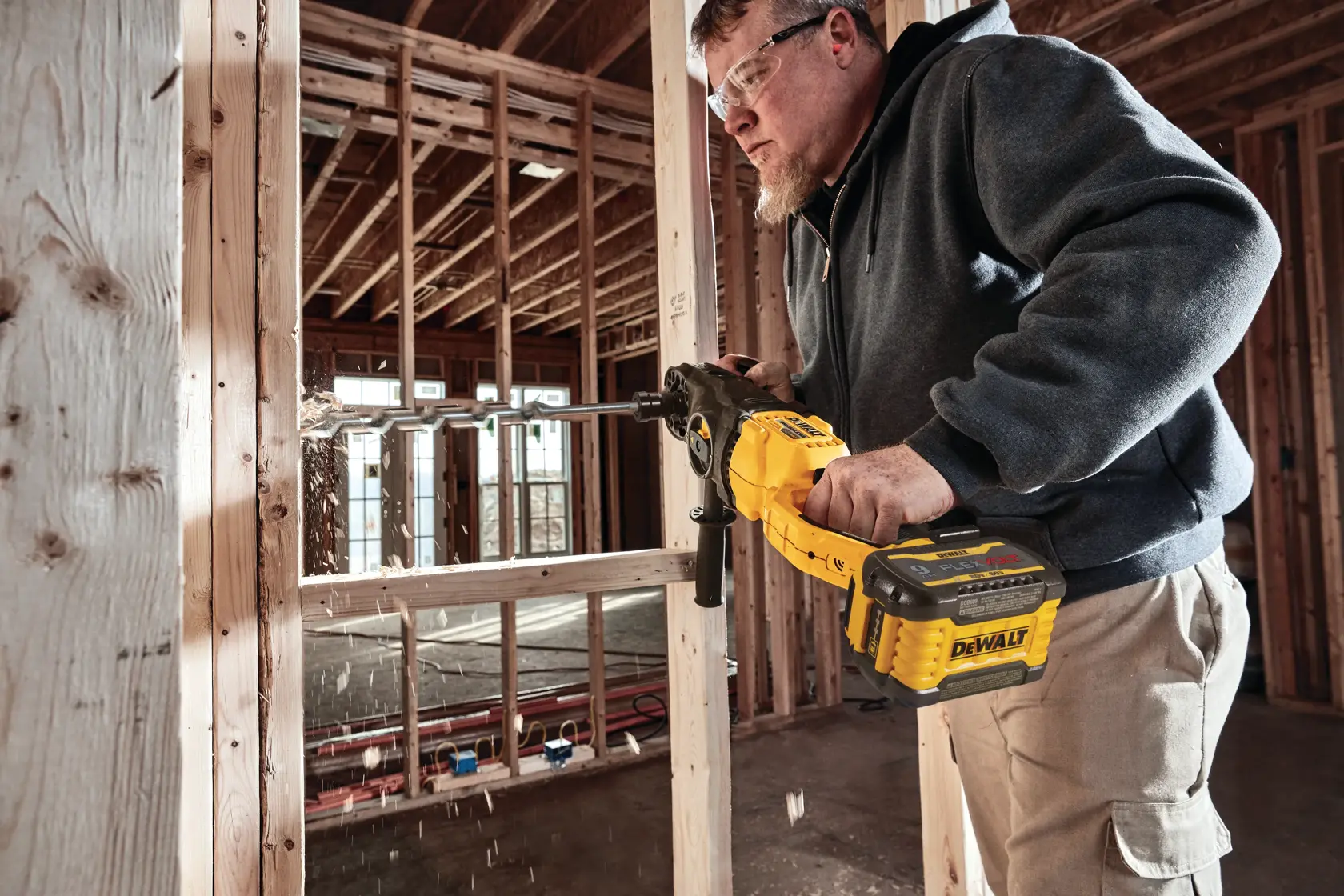 A person using a DEWALT cordless drill to bore a hole in a wooden frame inside an unfinished building under construction. The person's face is obscured, and wood shavings are seen flying from the drilling process.