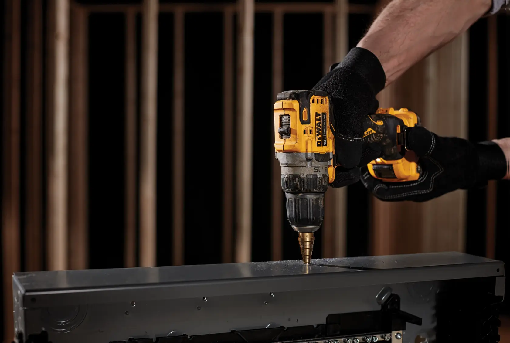 A person wearing black gloves is using a yellow DEWALT cordless drill to drill into a metal surface.
