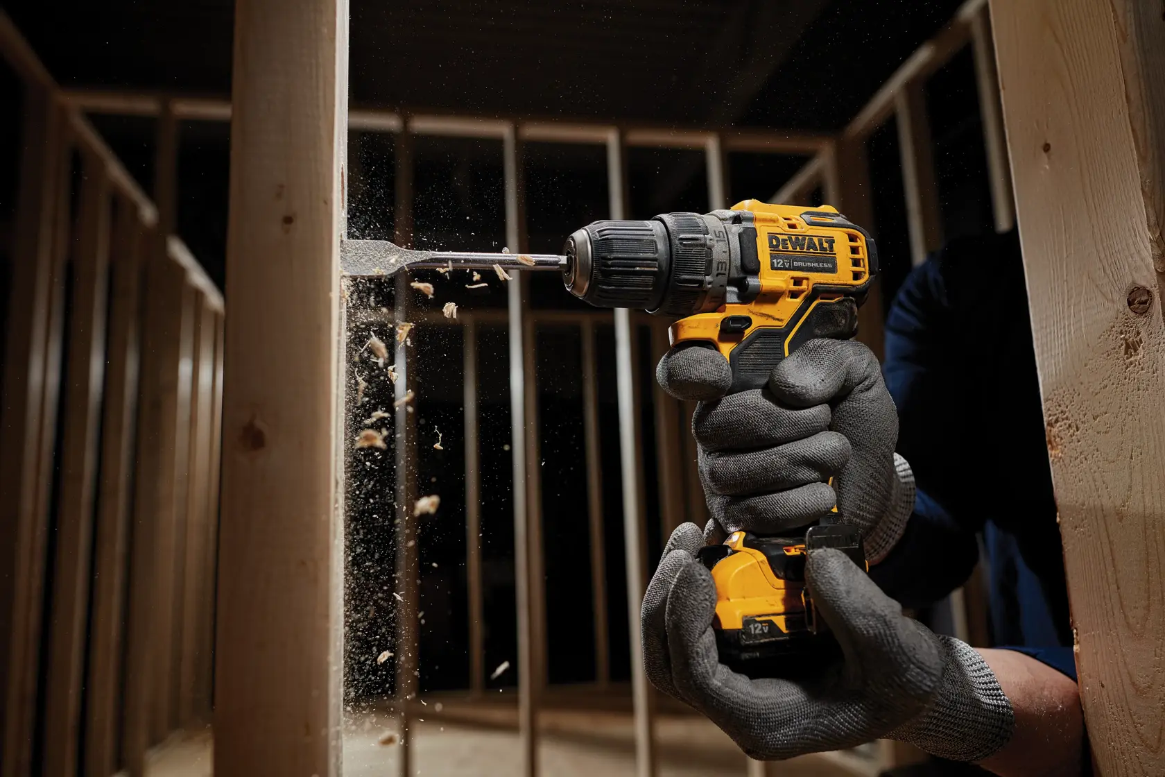 A person wearing gloves uses a yellow DEWALT cordless drill to bore a hole into a wooden beam. Sawdust and wood particles are visible as the drill works. The scene takes place in a construction environment with wooden framing.
