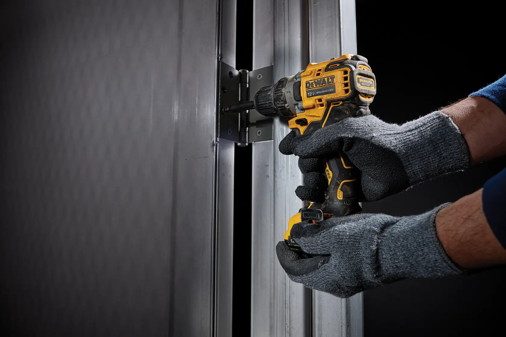 A person wearing gray gloves uses a yellow and black DEWALT cordless power drill to install a metal hinge on a vertical metal frame.