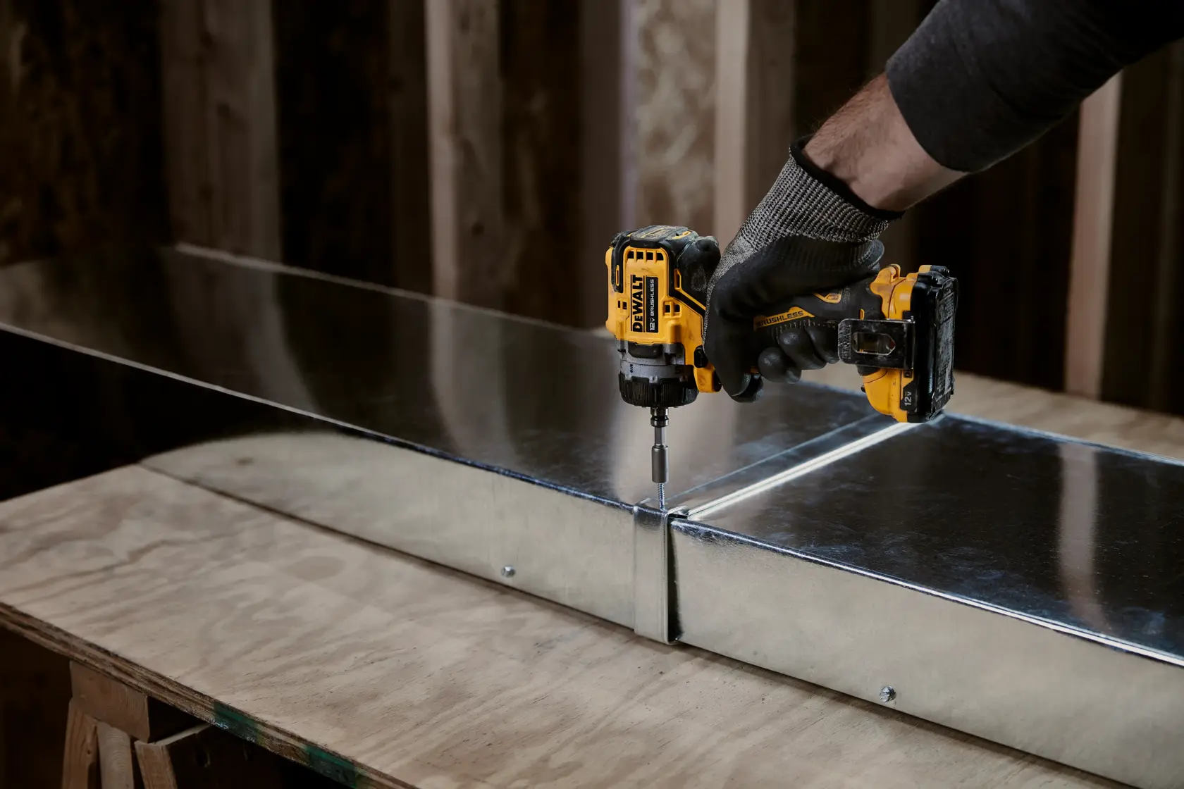 A person wearing a work glove uses a yellow and black DEWALT cordless drill to drive a screw into a piece of metal ductwork positioned on a wooden workbench.