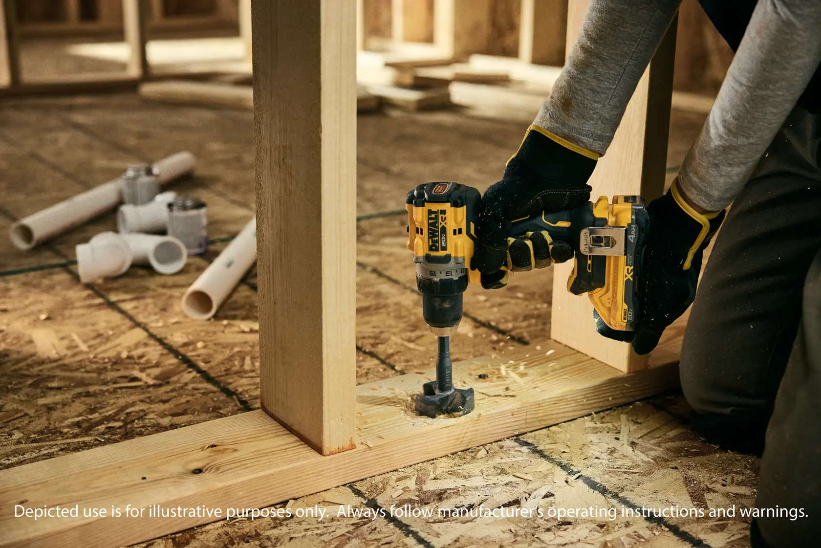 A person wearing gloves is using a DEWALT cordless drill to drill a hole in a wooden plank at a construction site. PVC pipes and fittings are visible on the floor in the background.