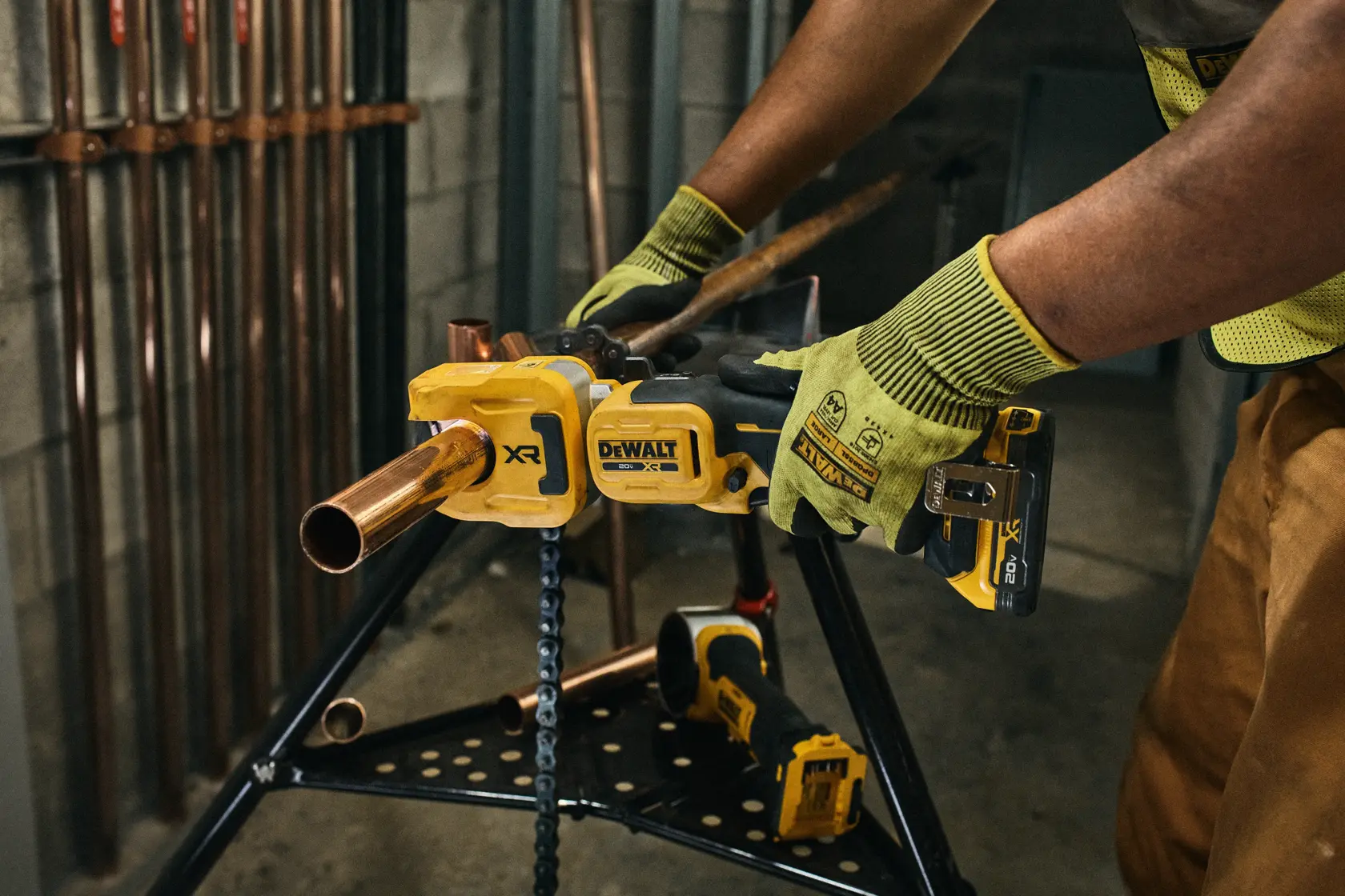 A person wearing work gloves is using a DEWALT cordless pipe cutter to cut a copper pipe, which is secured on a metal stand. The scene appears to be in an industrial or construction setting with copper pipes visible in the background.