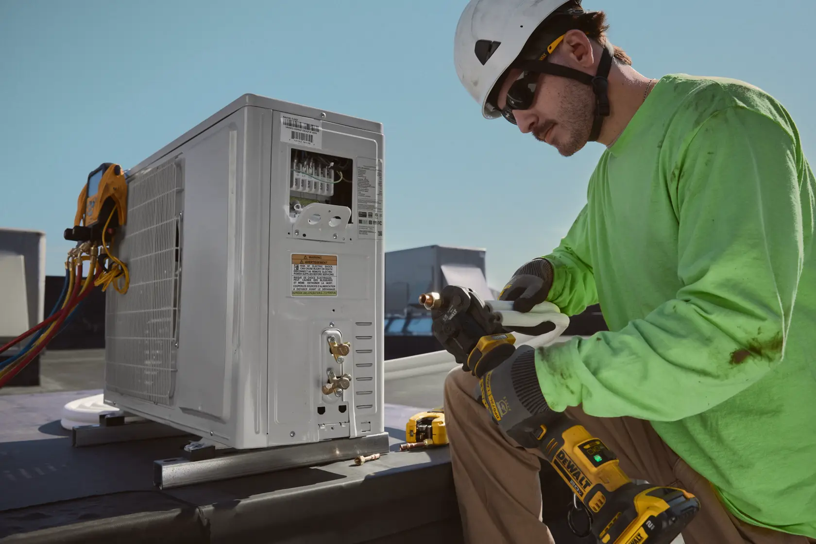 A person wearing a green shirt and hard hat uses a DEWALT cordless power tool to work on a HVAC unit on a rooftop. The unit has cables and meters attached, and the DEWALT tool is being used to connect or service piping.