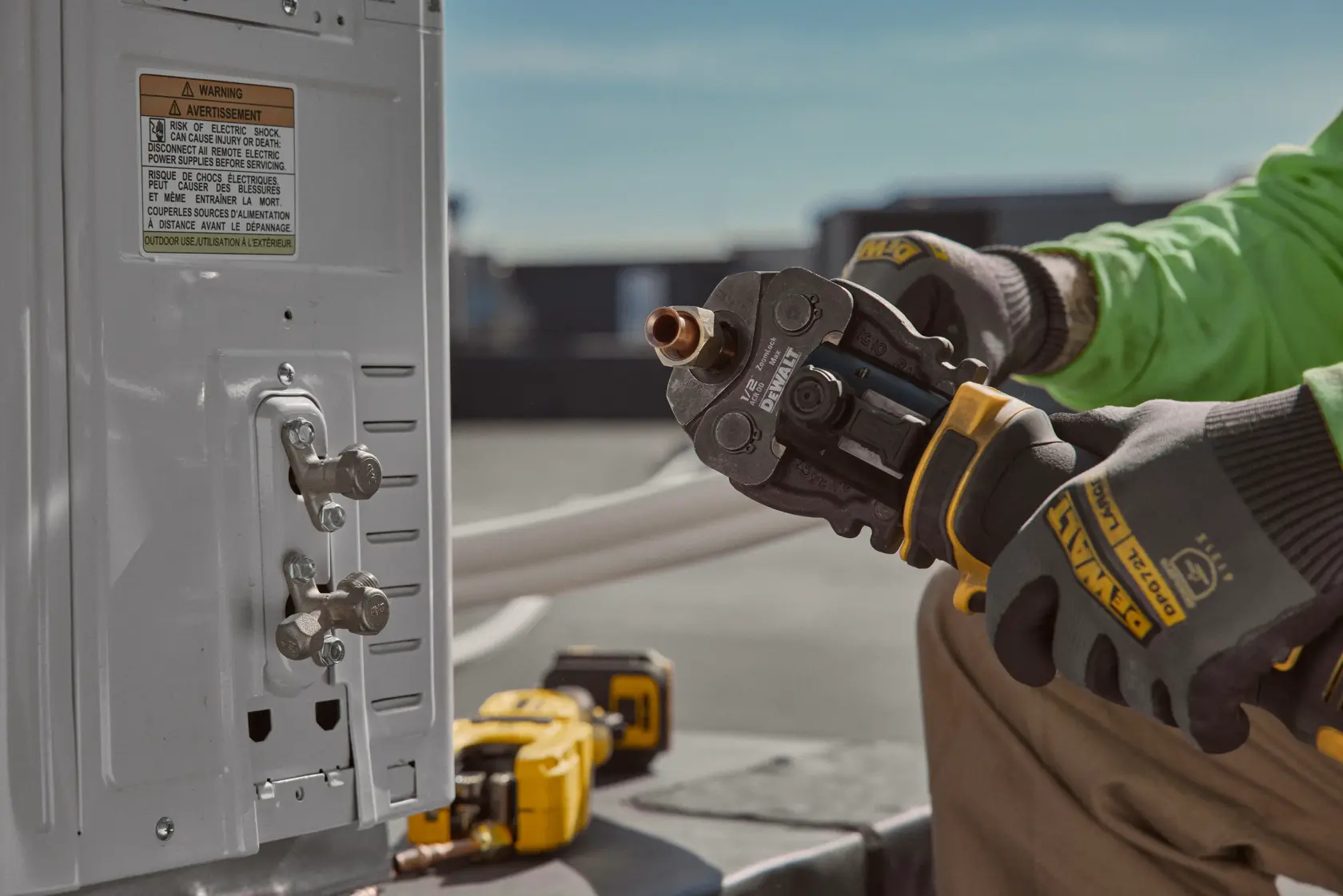 A person wearing DEWALT gloves is using a DEWALT power tool to work on a copper pipe attached to an electrical unit outdoors.