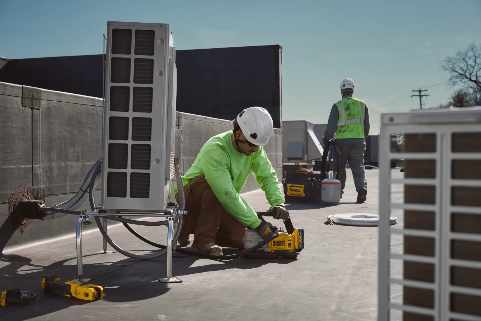 Two workers in safety gear, including hard hats and gloves, are working on a rooftop next to HVAC units. One worker is kneeling and using a yellow DEWALT tool, while the other is standing near additional equipment.