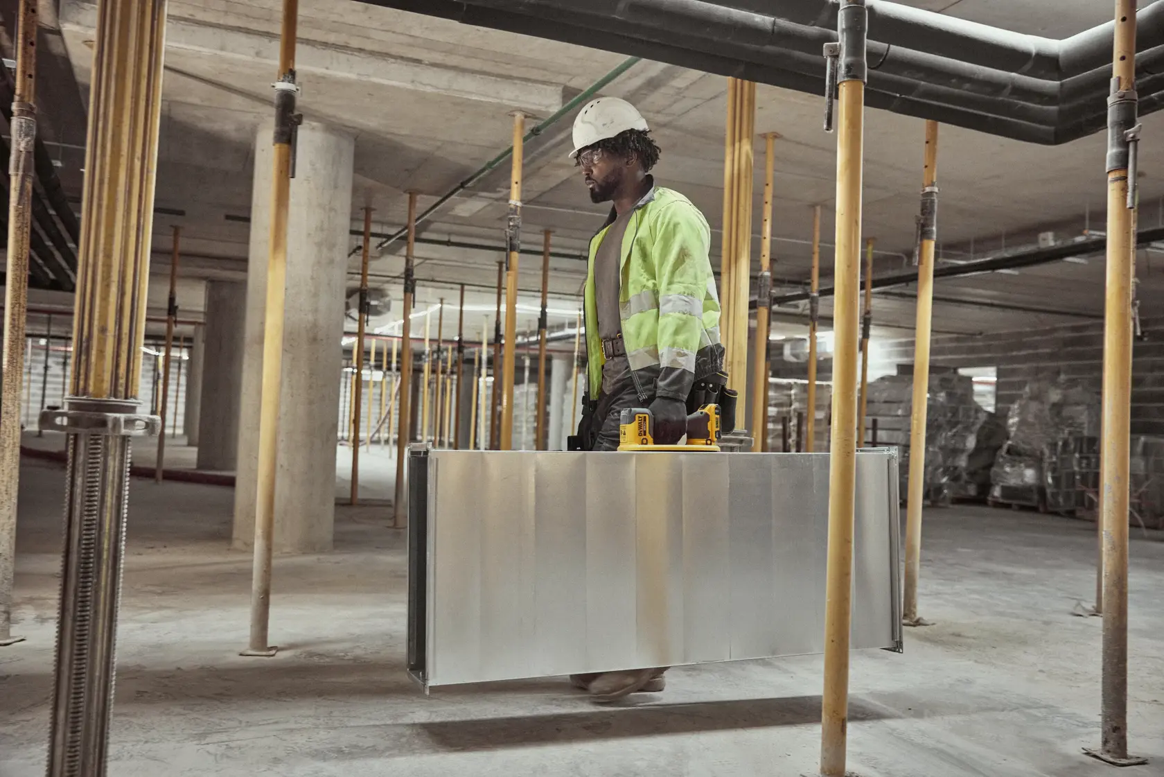 A person wearing a high-visibility jacket, hard hat, and gloves is carrying a large metallic panel with a DEWALT yellow power tool attached, inside a construction site with structural supports and unfinished surfaces.