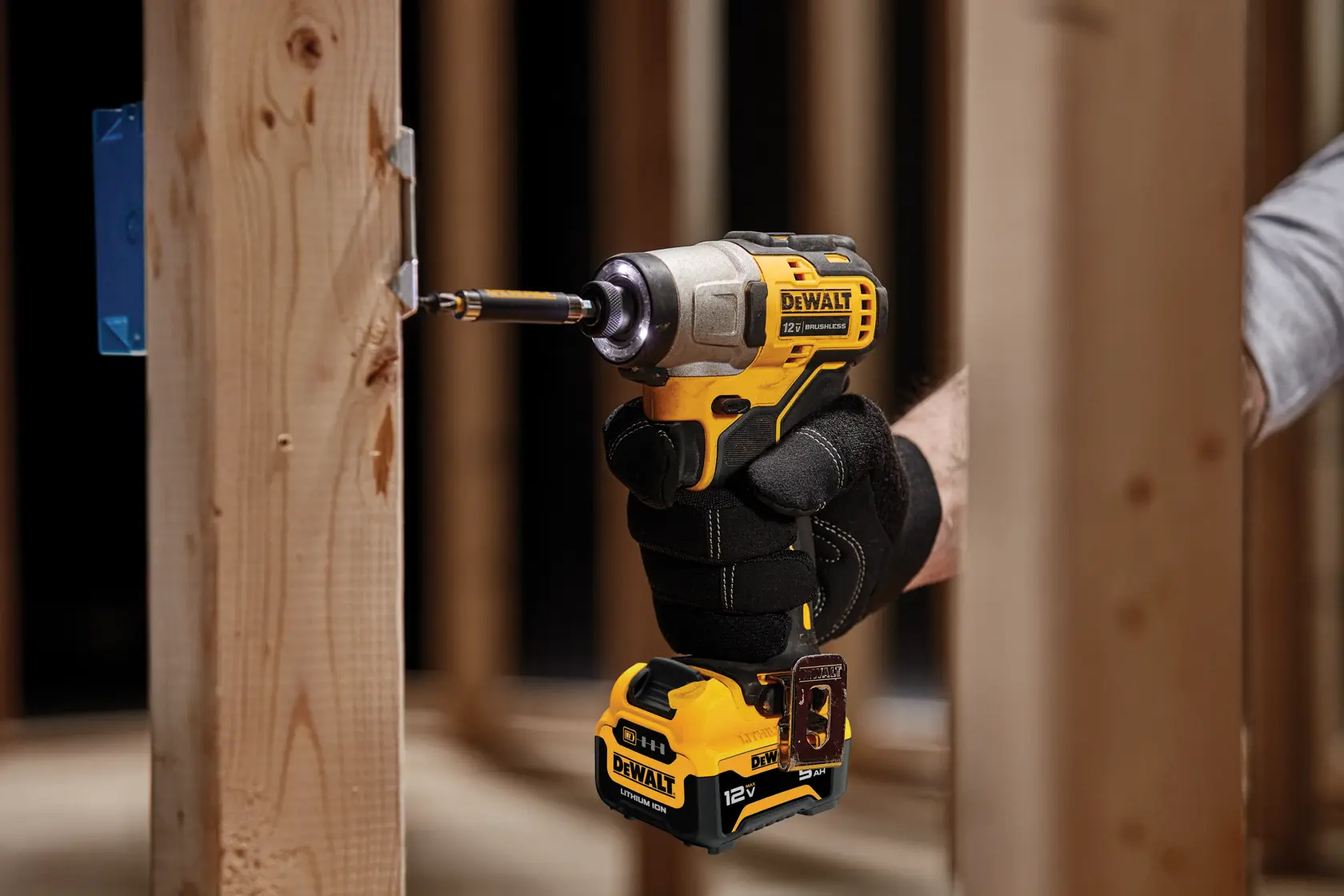 A close-up of a DEWALT DCF801J1 cordless impact driver being used to drive a screw into a wooden frame. The tool is held by a gloved hand in a construction setting.