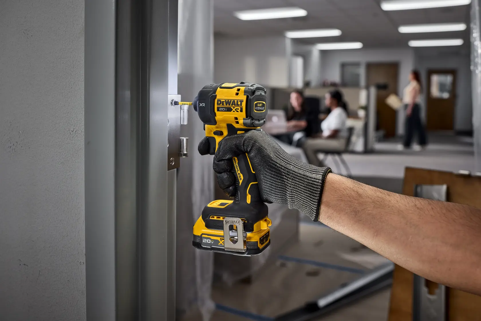 A hand wearing a work glove is using a DEWALT cordless impact wrench to install a metal latch on a door in an office setting. The DEWALT tool is yellow and black, and the background shows people and office furniture.