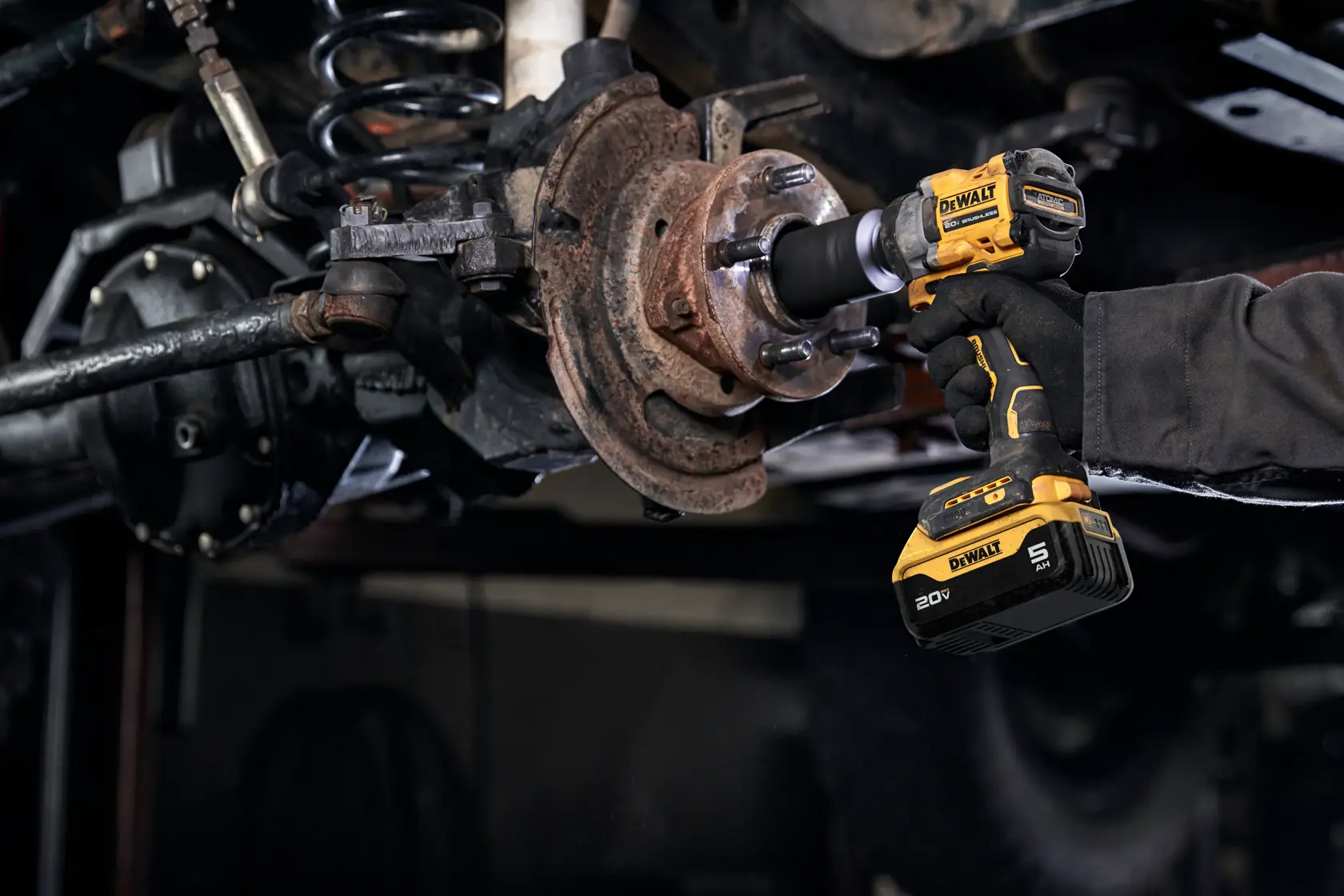 A person using a yellow and black cordless power tool on a vehicle's rusty wheel hub assembly, possibly for repair or maintenance.