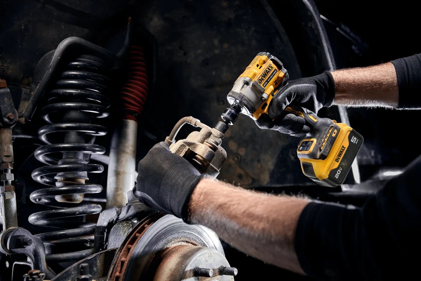 A person wearing black gloves uses a yellow cordless impact driver to work on a car suspension and brake assembly.