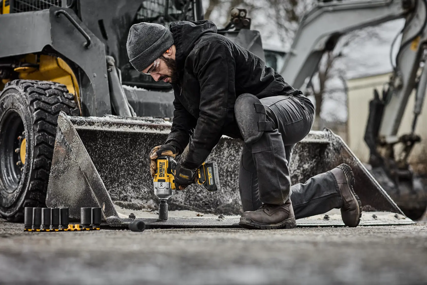 A person kneeling outdoors is using a DEWALT cordless impact wrench to work on a construction vehicle attachment. Several socket attachments are placed on the ground nearby. The person is dressed in dark work clothing and the scene appears to be in a cold environment.