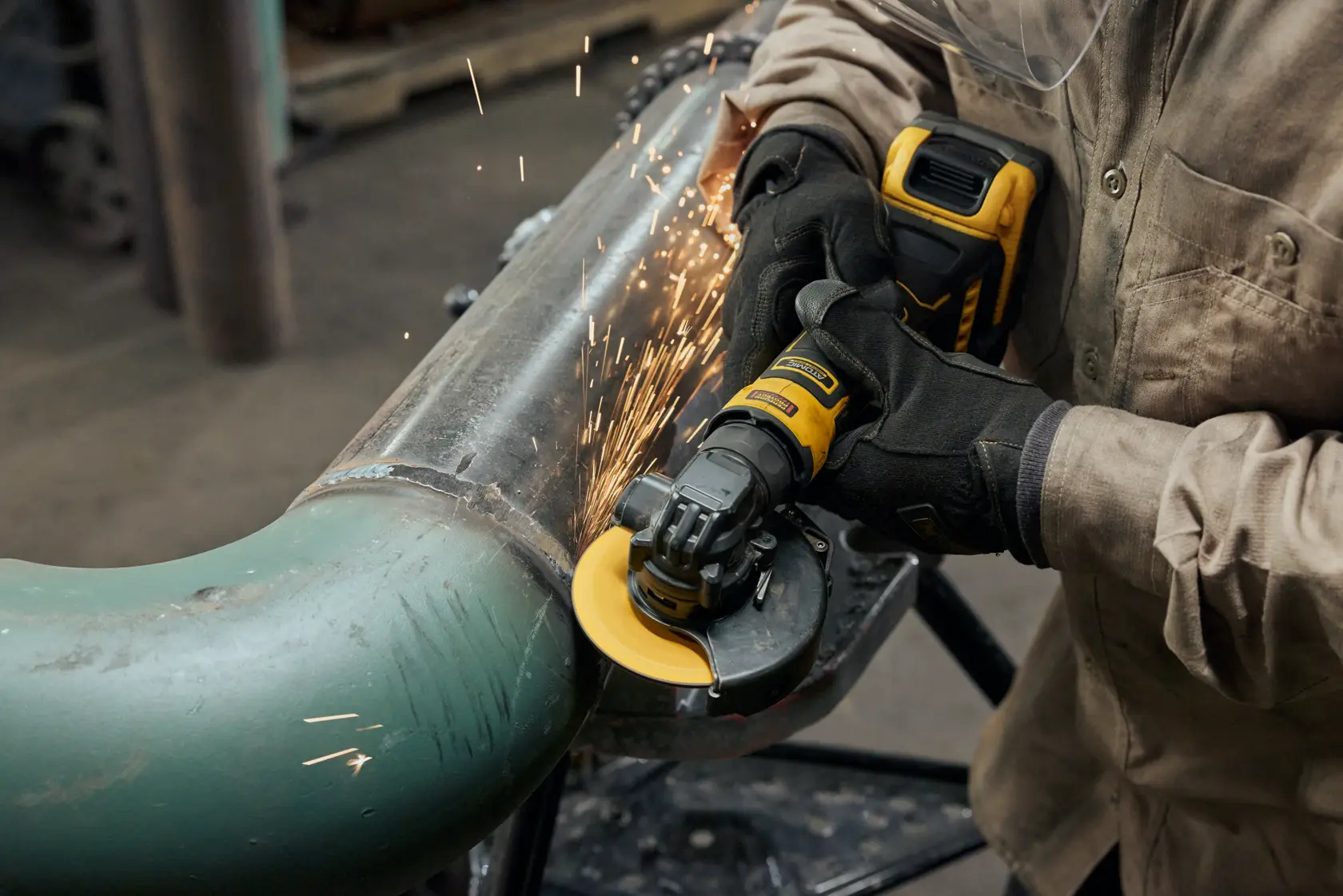 A person wearing protective gloves and clothing uses a DEWALT cordless angle grinder to cut or grind a metal pipe, with sparks flying from the operation.