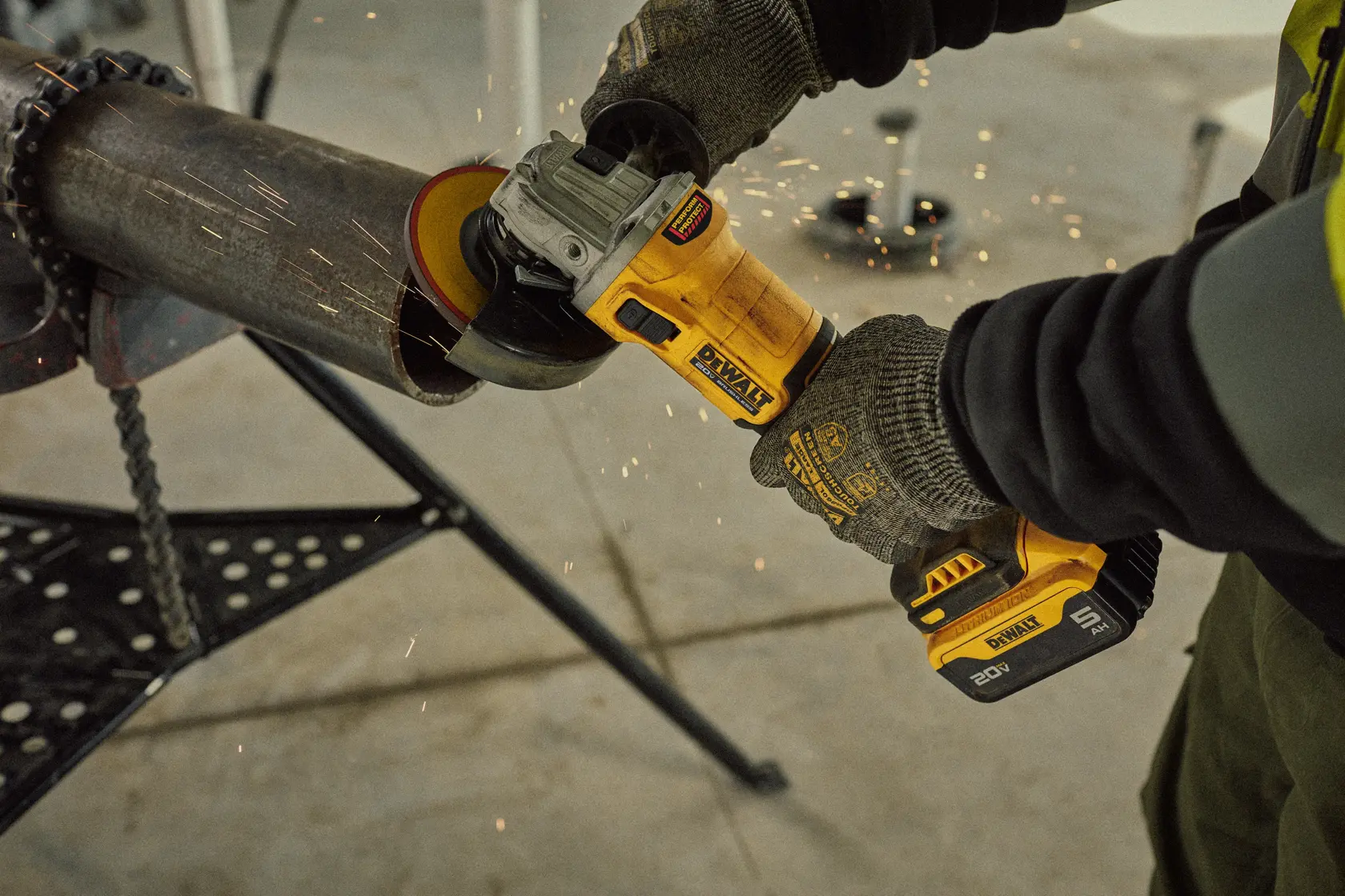 A person wearing gloves uses a DEWALT cordless angle grinder to cut or grind a metal pipe, with sparks flying from the cutting action. The tool is yellow and black and operated with a 20V battery.