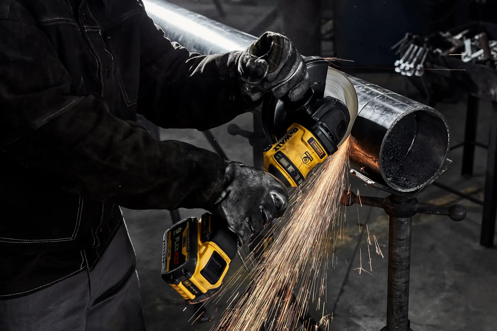 A person wearing protective gloves uses a yellow DEWALT cordless angle grinder to cut a metal pipe, with sparks flying from the cutting area.