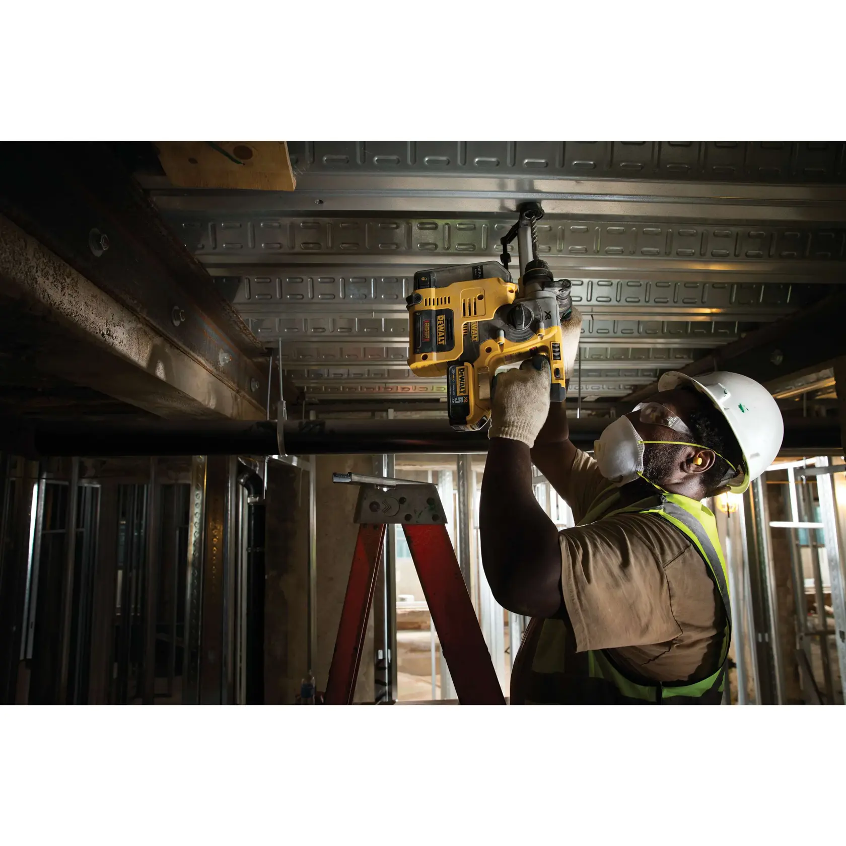 A construction worker wearing a safety helmet, high-visibility vest, gloves, and a face mask is using a yellow DEWALT rotary hammer drill (model DCH273P2) to drill into a metal ceiling, standing on a red ladder inside an unfinished industrial building.