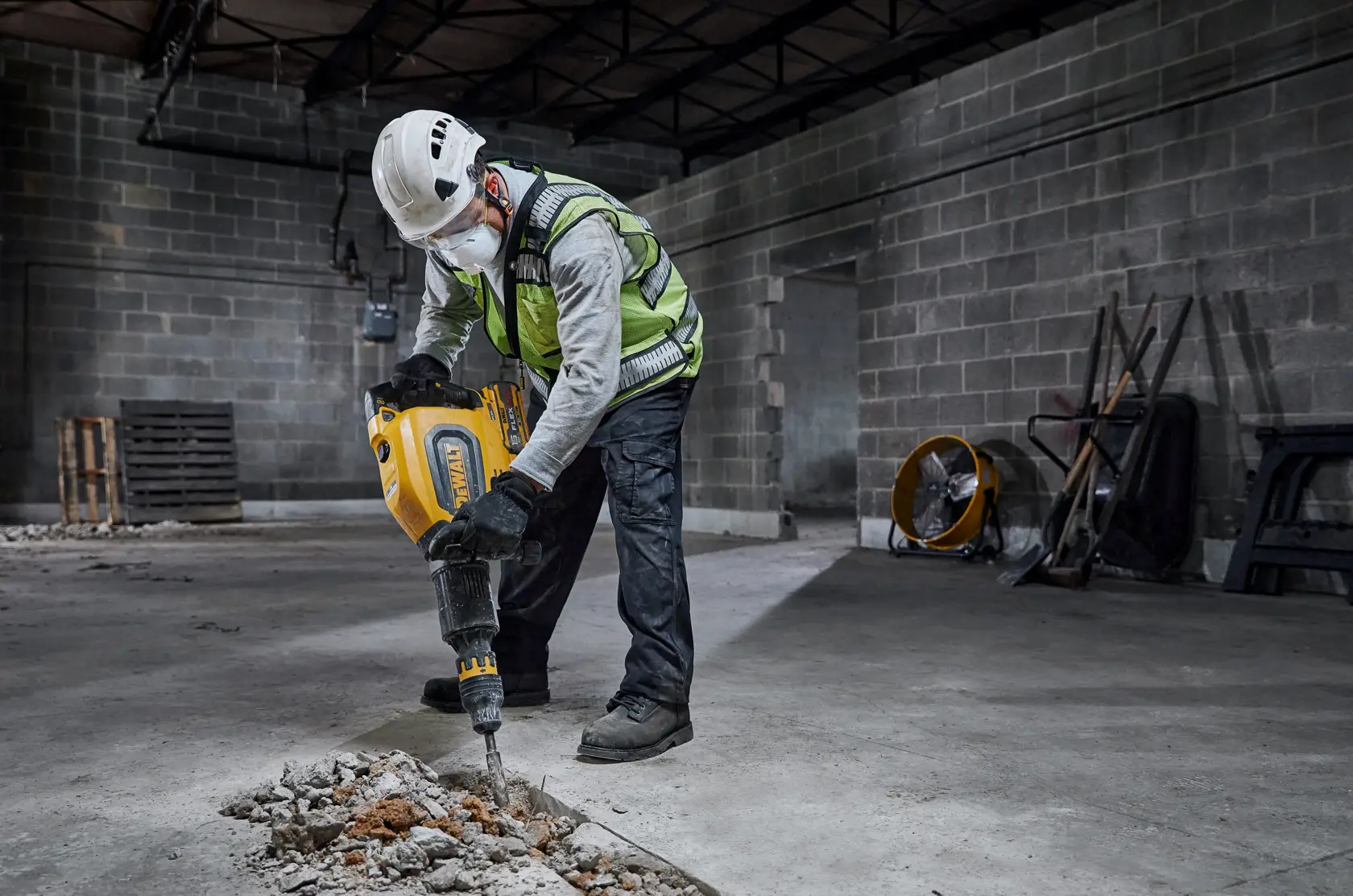 A person wearing safety gear uses a yellow DEWALT demolition hammer to break concrete inside an industrial building.
