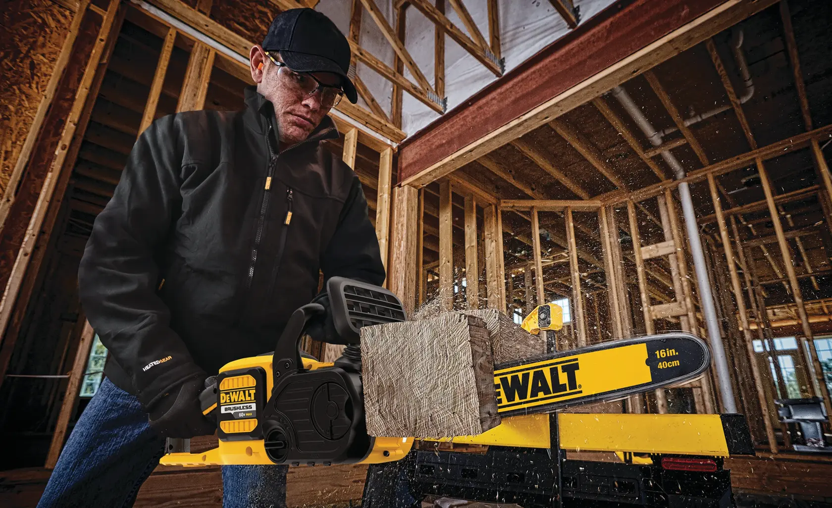 A person wearing a black jacket and dark hat is using a yellow DEWALT 16-inch (40 cm) electric chainsaw to cut through a piece of wood inside a building under construction with visible wooden framing.