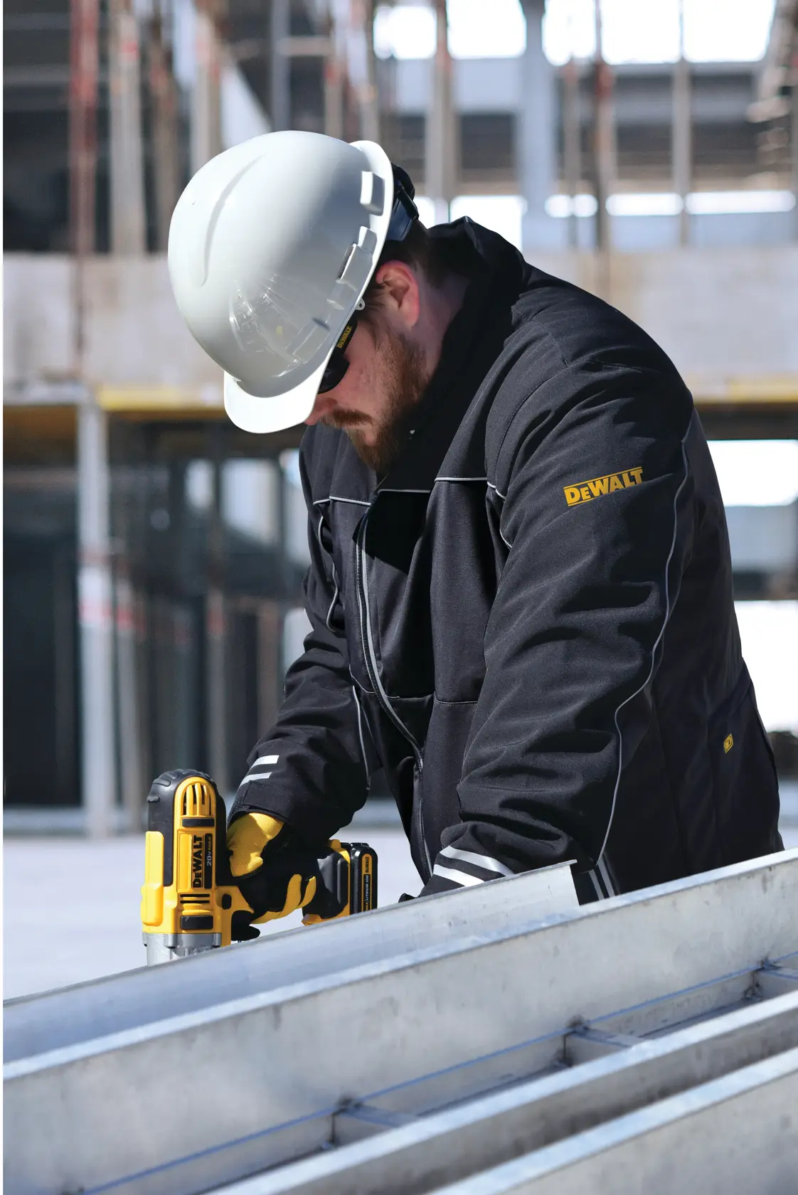 A person wearing a white safety helmet and a black DEWALT jacket is using a yellow and black DEWALT power tool at a construction site. The person's face is blurred for privacy.