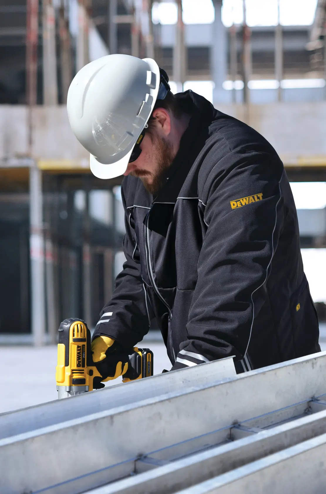 A person wearing a DEWALT branded black jacket and a white safety helmet is using a DEWALT power tool at a construction site. The jacket and tool are both visibly branded, and the surroundings include unfinished building structures.