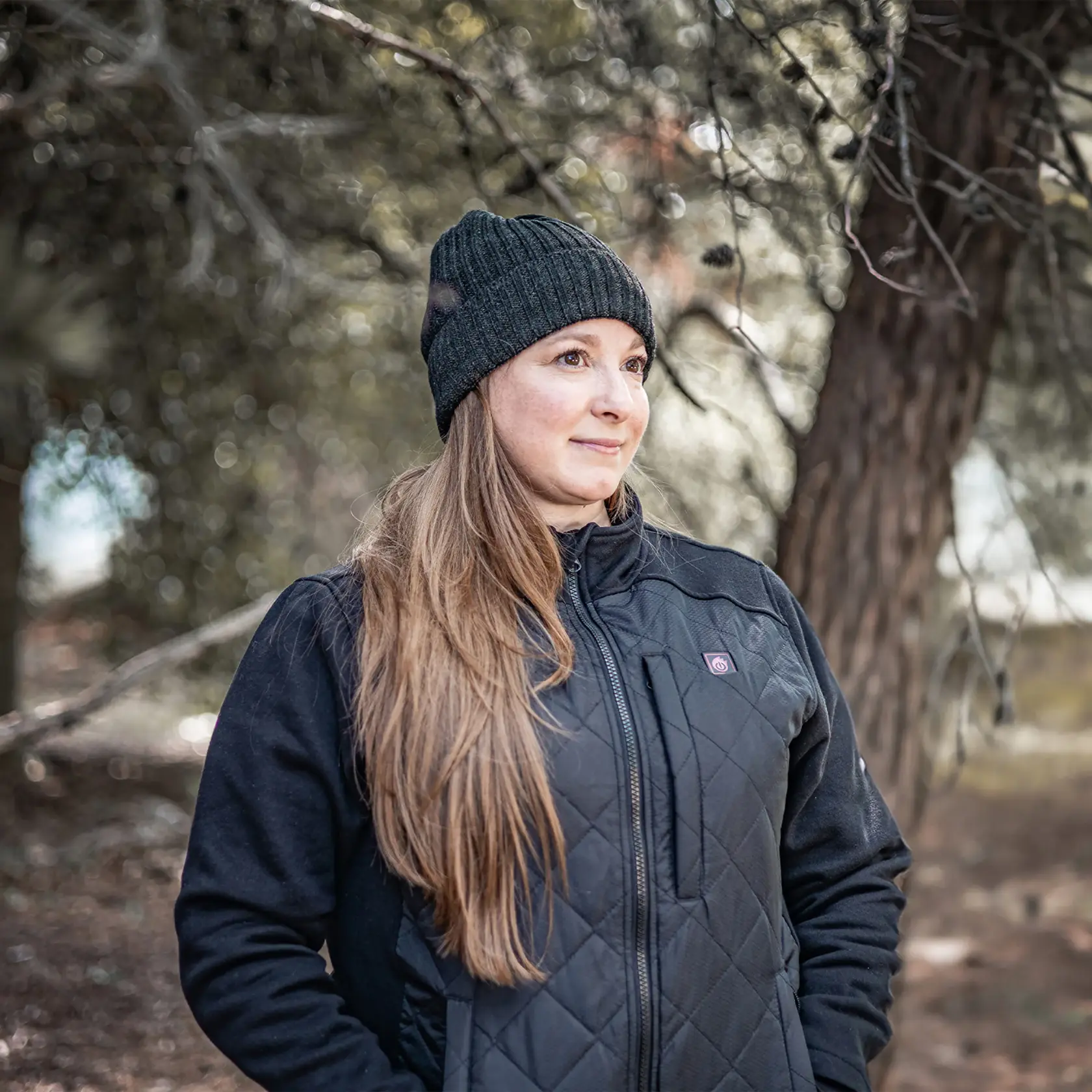 A person with their face blurred is wearing a black quilted DEWALT heated jacket and a black knit beanie, standing outdoors in a wooded area.