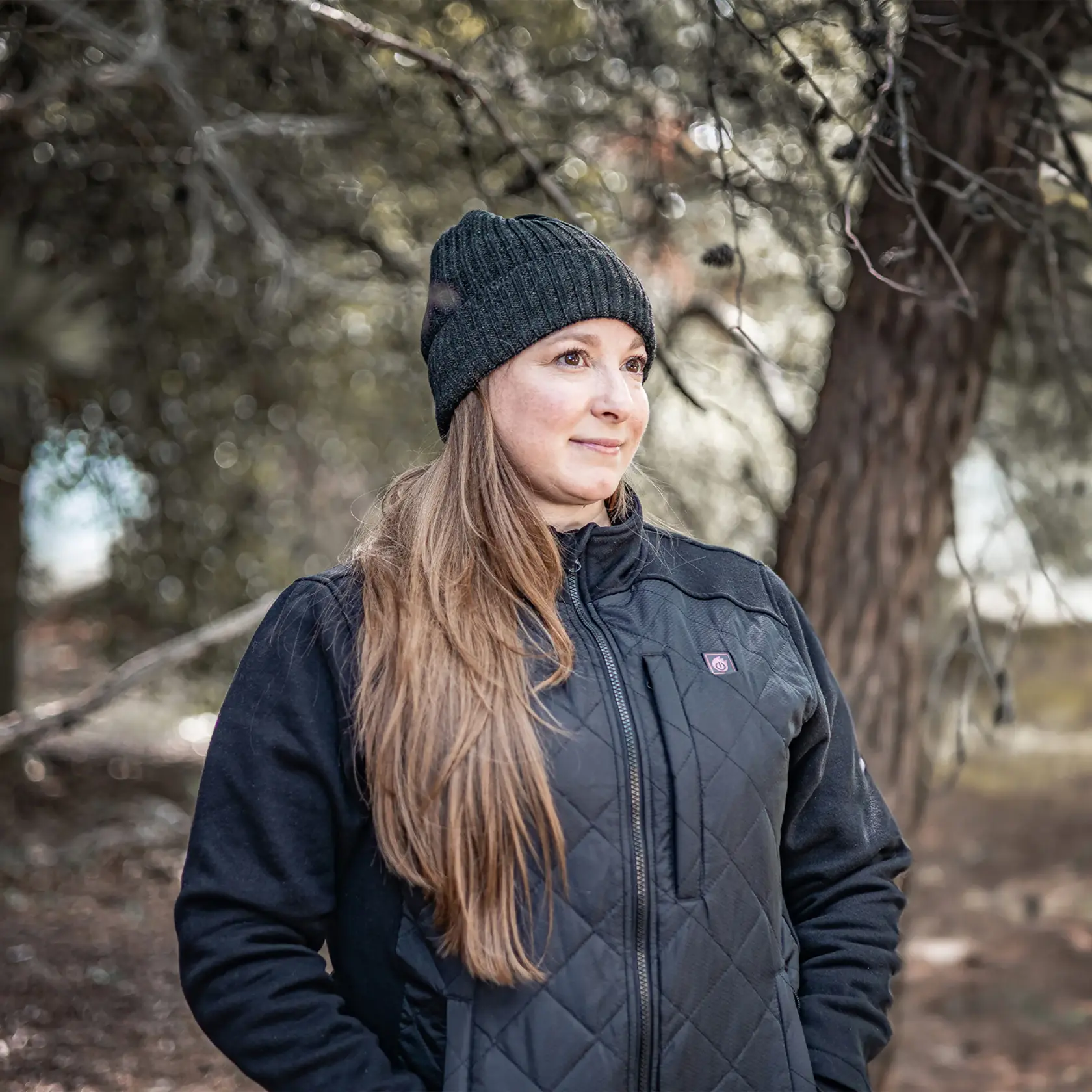 A person outdoors wearing a black quilted DEWALT heated jacket with a front zipper and a black knitted hat. The face is intentionally blurred for privacy, and trees are visible in the background.