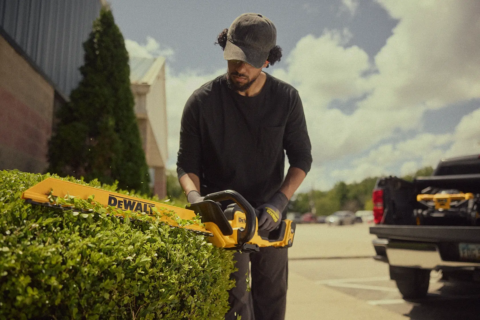 A person wearing gloves and a cap is trimming a hedge with a DEWALT cordless hedge trimmer outdoors near a parking lot. The trimmer is bright yellow and black.