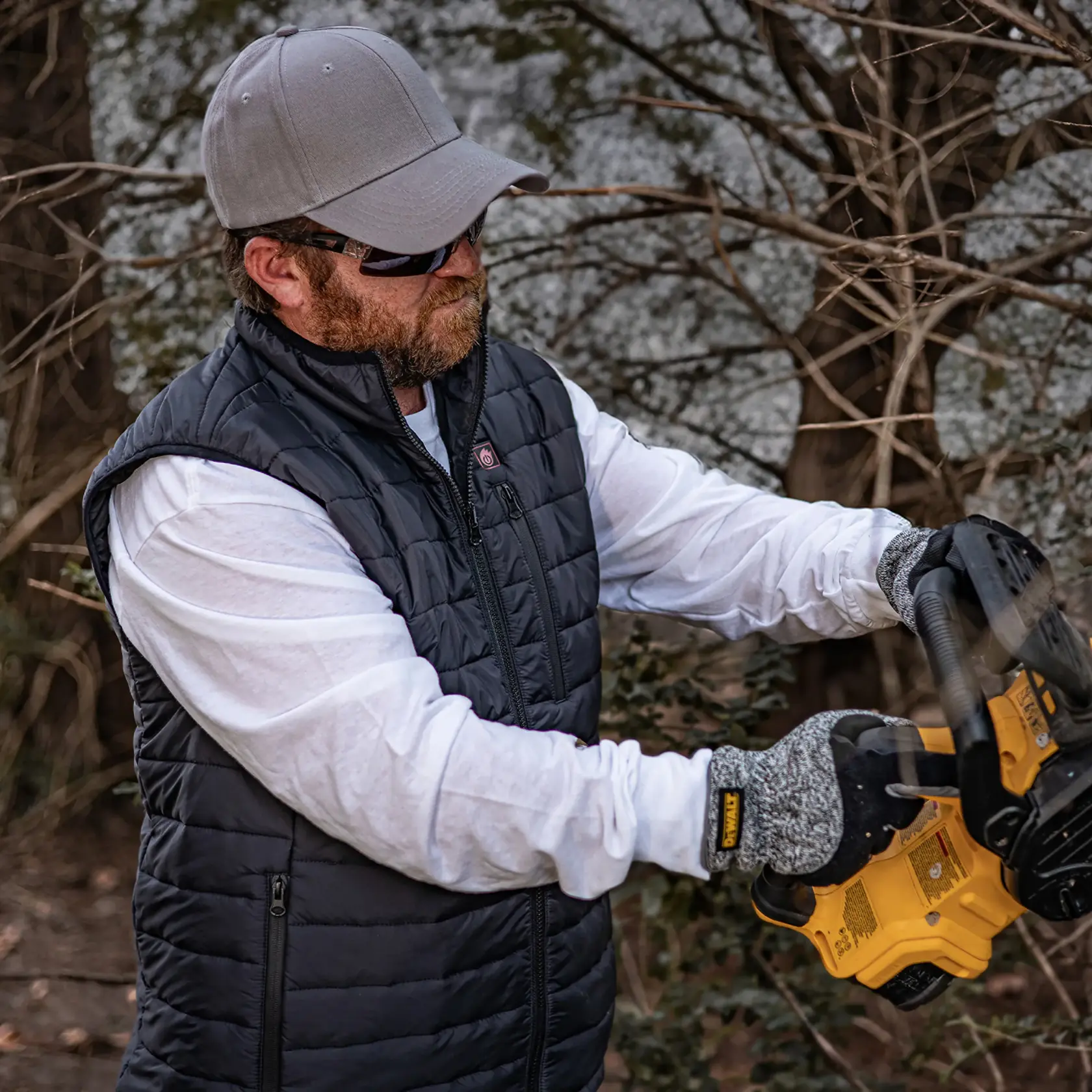 A person wearing a gray cap, black insulated vest, white shirt, and gray gloves is using a yellow DEWALT power tool outdoors. Trees and branches are in the background.