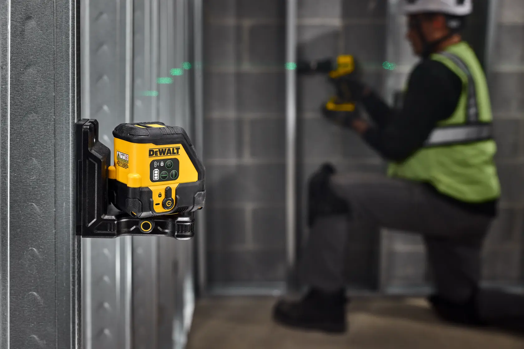 A yellow and black DEWALT laser level device is mounted on a metal frame wall, projecting a green laser line. In the background, a person wearing safety gear is kneeling and working on the metal structure.