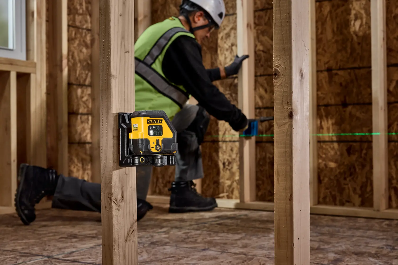 A DEWALT green beam laser level is mounted on a wooden stud in a construction area. In the background, a person wearing a safety vest, hard hat, and gloves is working on interior framing, with the laser casting a visible green line for alignment.