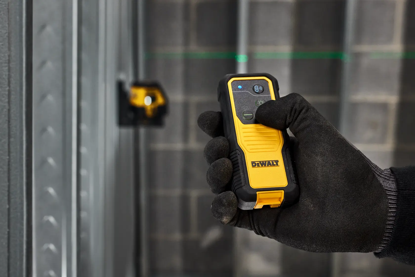 A gloved hand holding a DEWALT laser distance measurer in an industrial setting, with a green laser line visible on the wall and a laser level tool in the background.